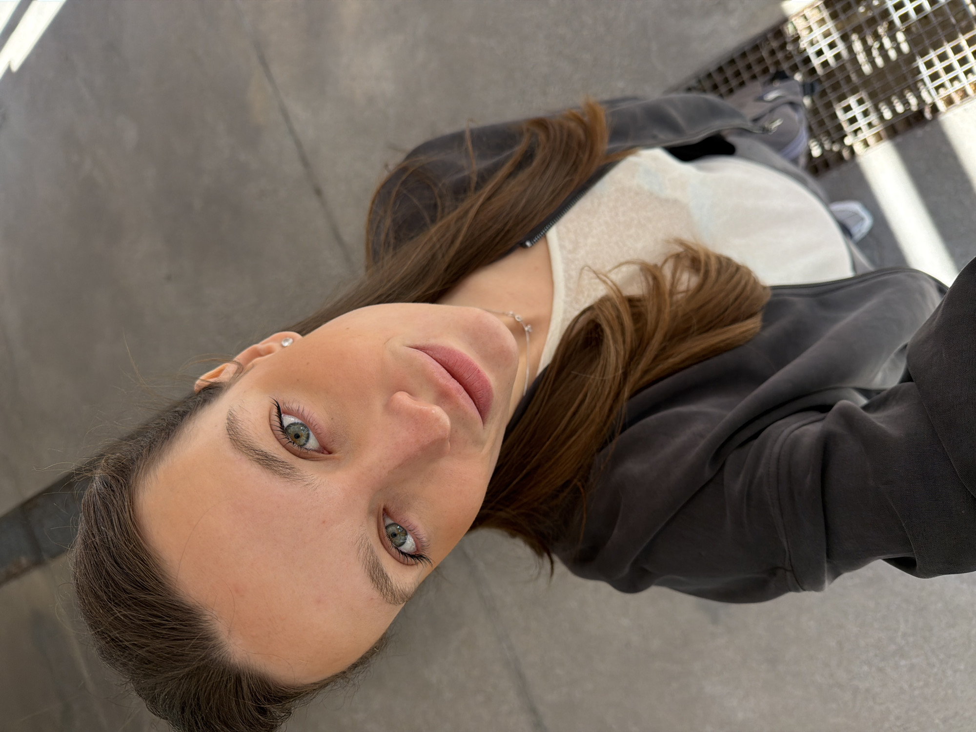 A woman with brown hair and green eyes takes a selfie at eye level, looking up towards the camera. She is wearing a gray jacket over a white top. The background consists of gray pavement, with a drain on the right side, suggestive of an urban setting. The photo may have been taken in Budva, Montenegro.