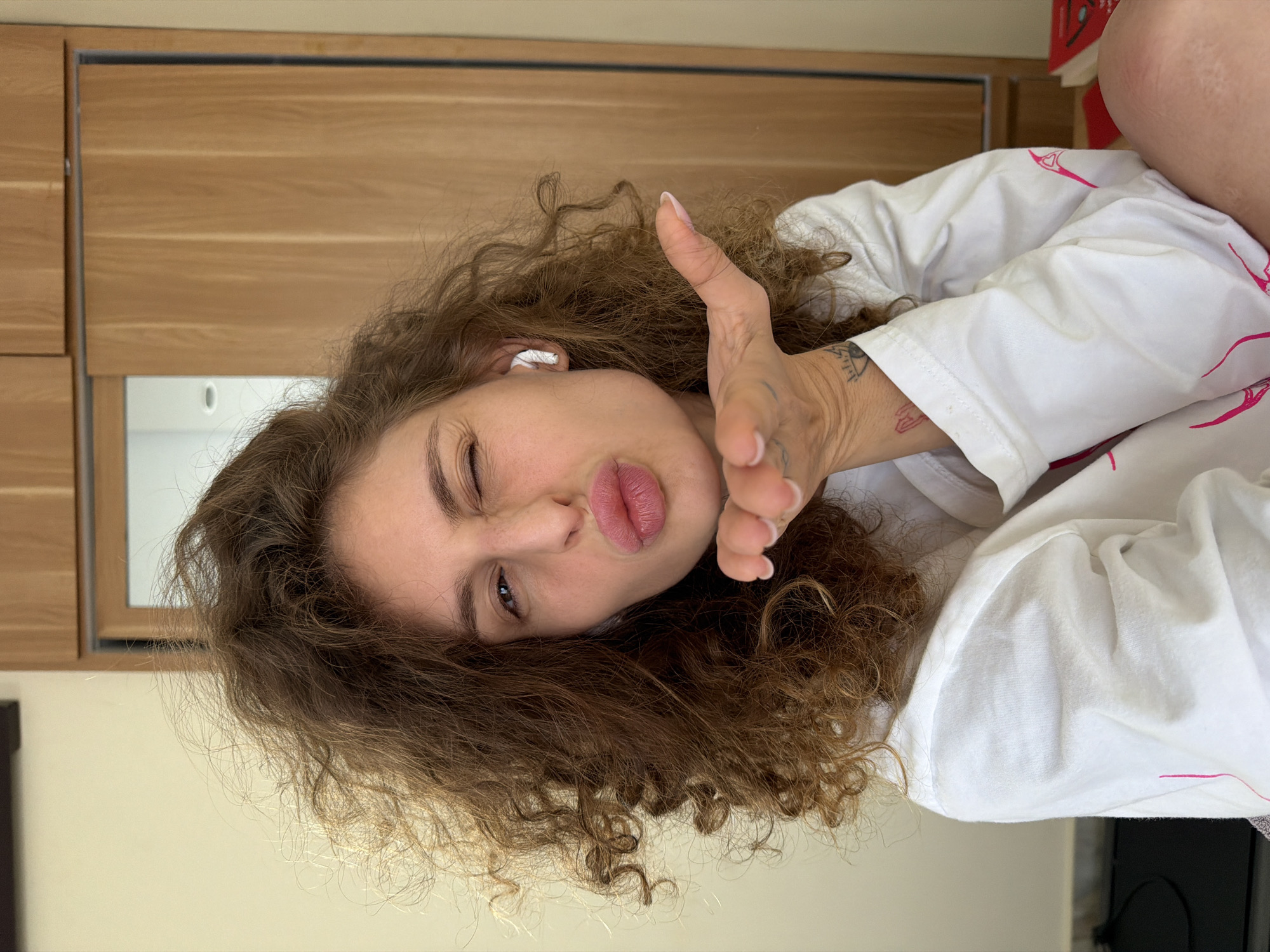 A light-skinned adult female, positioned in a medium close-up, is depicted indoors in North Kuta, Indonesia. Her brown, curly hair is voluminous and extends around her head. She is winking with her right eye and puckering her lips in a kissing gesture, with her left hand raised towards the camera, palm up, forming a shape as if blowing a kiss. A white wireless earbud is visible in her right ear. She wears a white, long-sleeved garment with faint pink patterns. Tattoos are present on her left wrist, including an eye symbol and a smaller, red design. The background features a plain, light-colored wall on the left and a wooden cabinet or sliding door unit on the right. A partial reflection of an interior space with a light fixture is visible in a mirrored section of the wooden unit. Red objects, possibly books, are partially visible on the far right.