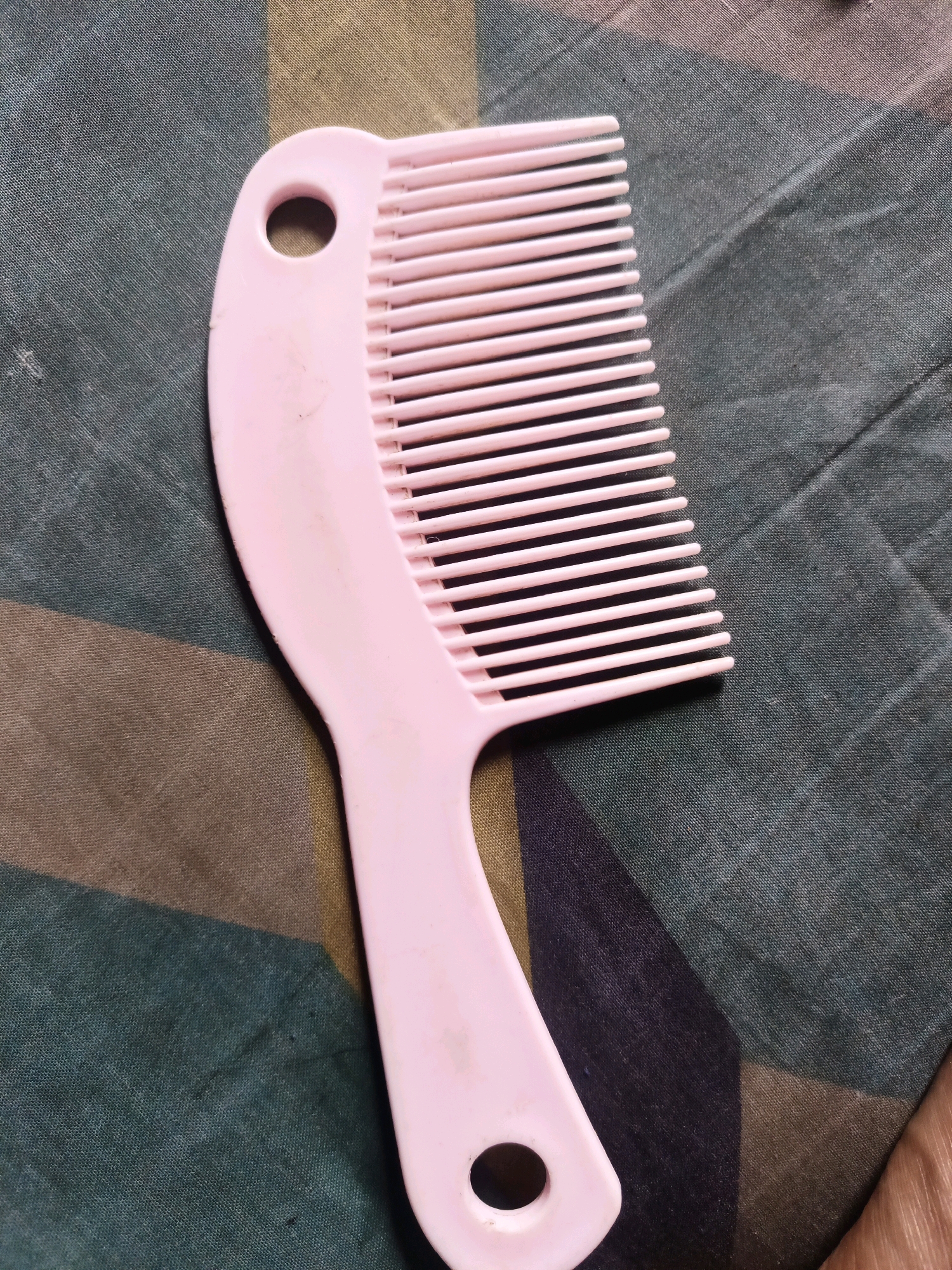 A pink plastic comb lies diagonally on a fabric surface. The fabric has a pattern of dark green, brown, and black stripes. The comb has a curved handle with a circular hole at the top and another at the bottom. The teeth of the comb are thin and evenly spaced. The image appears to have been taken from above. There is no indication of the location in South Jakarta, Indonesia, or any people, actions, or interactions.