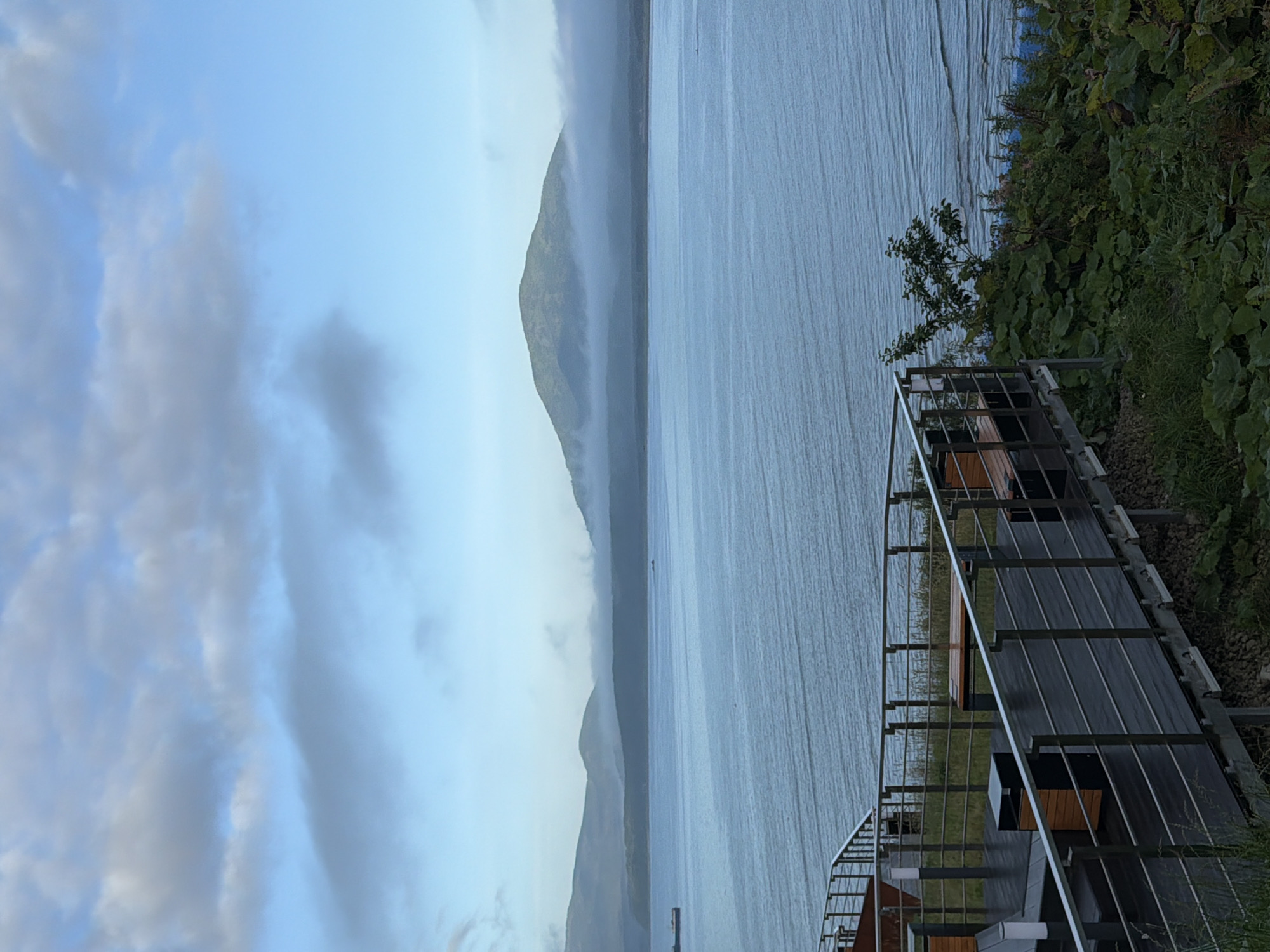 An elevated wooden deck with benches and trash receptacles overlooks a calm, blue body of water. A metal railing lines the edge of the deck. In the distance, mist partially obscures rolling green hills and a prominent, rounded mountain. The sky above is a pale blue with scattered, light gray clouds. A ship is visible on the water to the left. The deck is situated on a grassy incline. The scene is likely a viewpoint or recreational area in Yuzhno-Kurilsk, Russia, given the landscape.