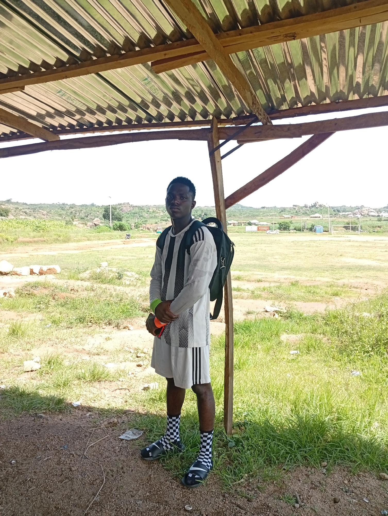 A young Black male stands under a corrugated metal and wooden shelter, facing the camera. He is wearing a grey long-sleeved shirt with black stripes, grey shorts with black stripes, black and white checkered socks, and black sandals. He has a black backpack slung over his left shoulder and is holding a bright orange object in his right hand, wearing a green wristband on his left wrist. The background shows a dry, grassy field with scattered rocks and some distant hills and buildings, consistent with a semi-arid landscape. This scene is likely located in Jos, Nigeria, known for its plateau landscape and rocky outcrops.