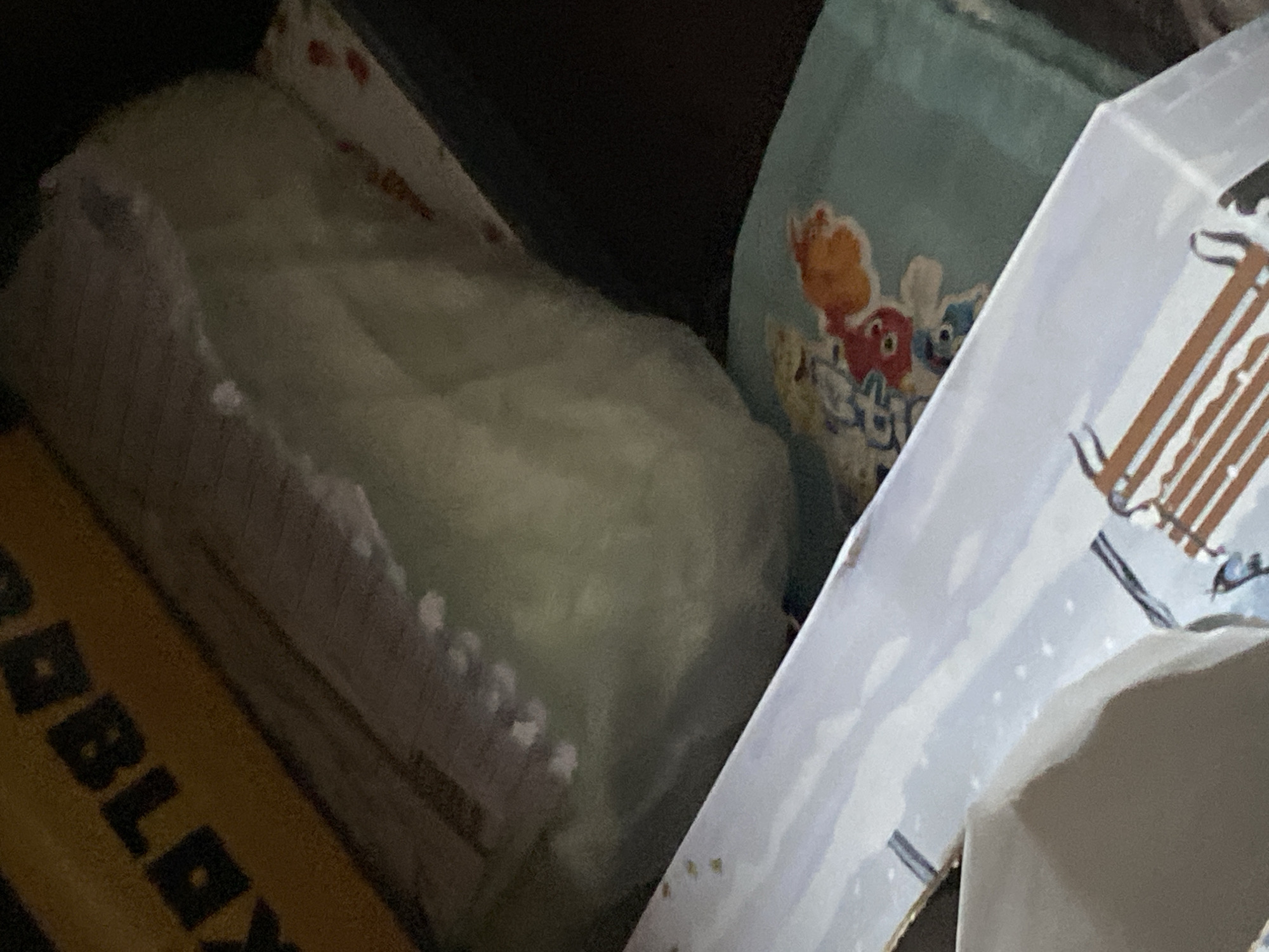 The image shows a close-up of a collection of items. A package with "ROBLOX" visible on it is in the lower left. Next to it is a package of soft goods that appears to be a stack of cloths or paper towels, and it has a red and white design on top of it. There is a bag with cartoonish animal figures printed on it. Finally, there is a white box or container with brown lines that resembles a building. The setting is indoors, and the light is not very bright. The location is unknown and no information in the image suggests it is in Sofia, Bulgaria.