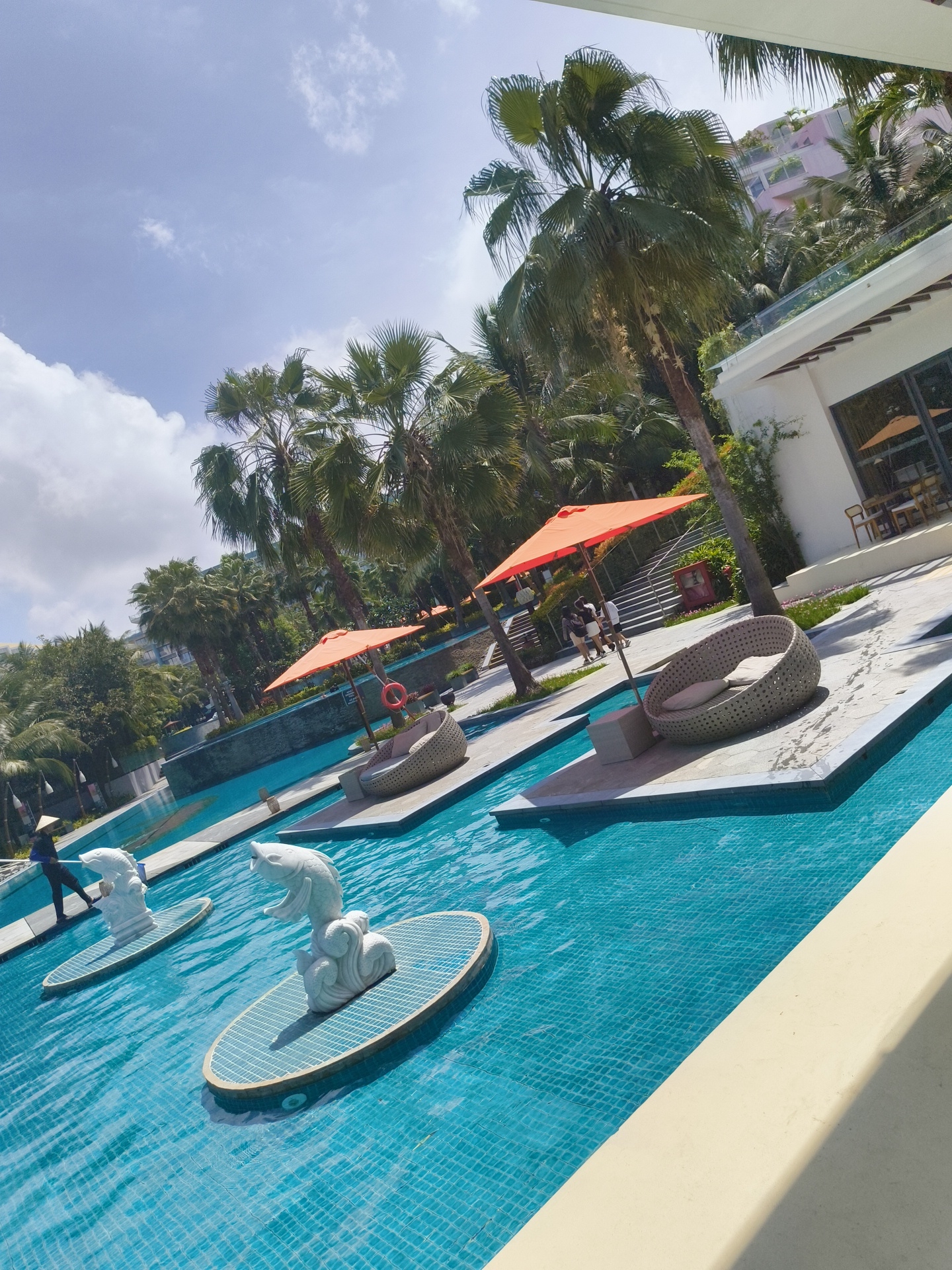 A resort swimming pool area in Phú Quốc, Vietnam, features a bright blue tiled pool with several floating platforms. On two of these platforms, white sculptures of leaping fish are positioned. A person wearing a conical hat and dark clothing is walking near the edge of the pool. Further in the pool, an orange umbrella shades a woven lounge chair on a concrete platform. Palm trees line the background, and a modern building with balconies is visible in the distance. The sky is blue with scattered white clouds.
