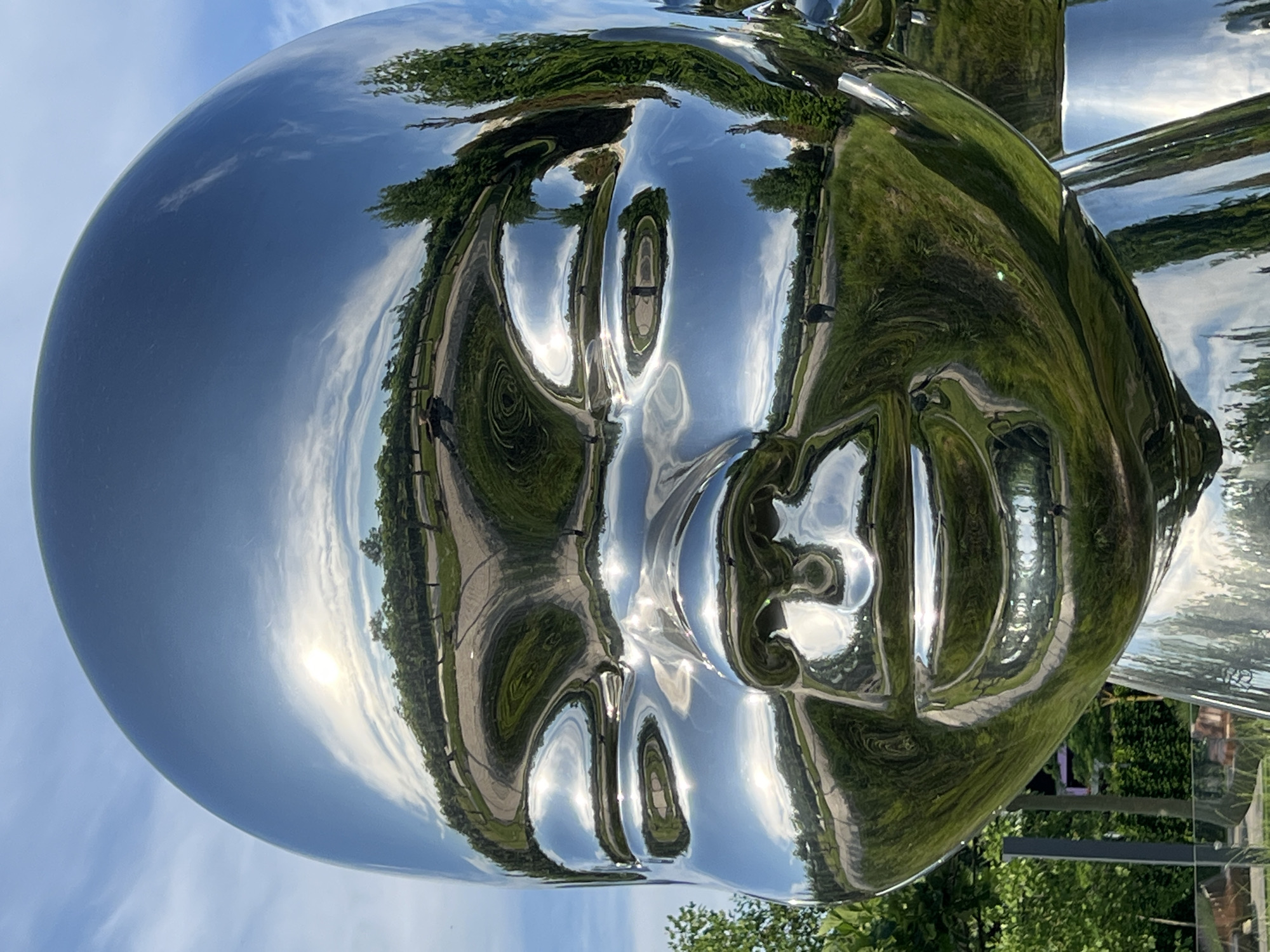 The image is a close-up, eye-level, wide-angle shot of a large, chrome-plated sculpture of a baby's head. The sculpture is highly reflective, mirroring the surrounding environment. A path, lined with trees, winds through a grassy area. The sun is visible in the sky, partially obscured by clouds. Several people are visible as reflections in the sculpture. The sculpture is located in the city of Krasnodar, Russia.