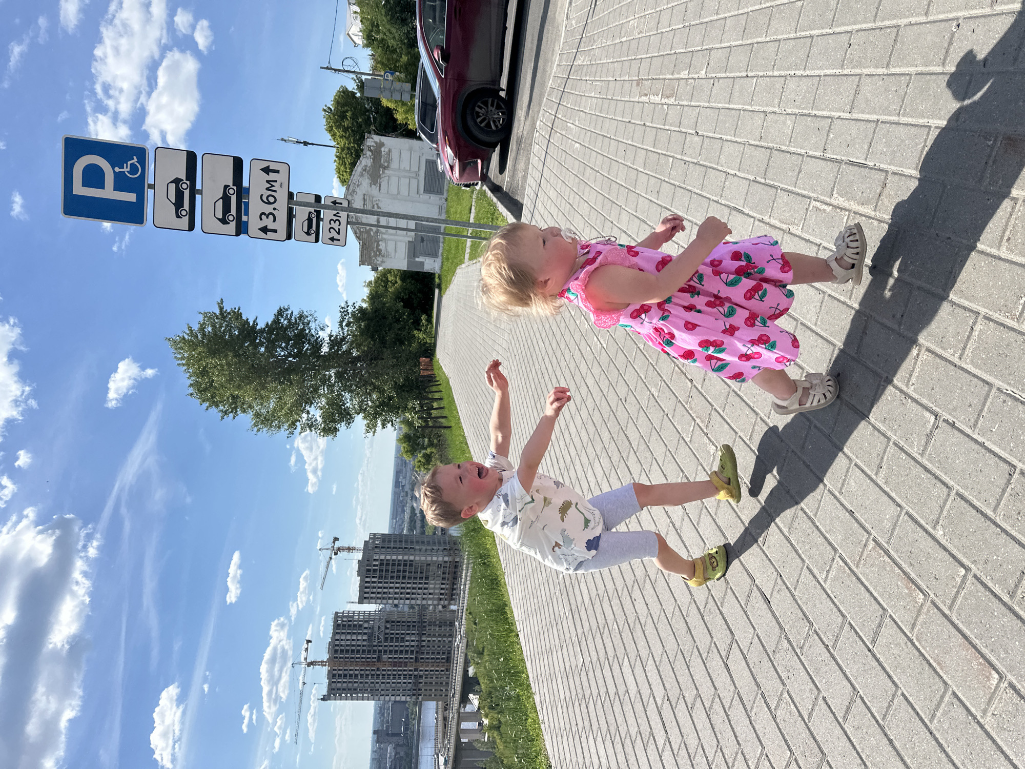The image depicts an outdoor, sunlit scene on a paved pedestrian walkway in Nizhny Novgorod, Russia, featuring two young children.
On the left, a young boy with light hair and fair skin is captured mid-stride, moving towards the right. He wears a white short-sleeved t-shirt with multiple light blue dinosaur illustrations, light grey shorts, and yellow open-toed sandals. His mouth is open, revealing an interior, and his cheeks appear flushed. His arms are extended forward at chest level, with hands open, suggesting an active pursuit or play. Behind him is a grassy incline.
To the boy's right and slightly ahead, a younger girl with light hair and fair skin walks to the right. She has a white pacifier in her mouth. She is dressed in a sleeveless pink dress adorned with repeating red cherry patterns and wears light beige open-toed sandals. Her gaze is directed slightly to her right.
The children are on a paved path composed of light grey rectangular pavers arranged in a herringbone pattern. Strong shadows are cast on the path by the children, indicating bright overhead sunlight.
In the mid-ground, a vibrant green grassy slope is visible behind the children, leading up to a line of trees. In the background on the far right, a tall metal pole displays a stack of five rectangular parking signs. The uppermost sign is blue with a white 'P' and a wheelchair symbol, indicating disabled parking. Below it are two signs featuring a black car silhouette. The fourth sign indicates "↑ 3,6M ↑" (3.6 meters height clearance), followed by another car silhouette sign, and then "↑ 23M ↑" (23 meters clearance/distance). Behind these signs, a red SUV, identified as a Lexus NX, is parked, partially obscured by trees and a light-colored building.
To the left of the background, a wide river is visible, spanned by a large concrete bridge featuring multiple lanes and a distinct arch structure on its left side. Beyond the river and bridge, two prominent, multi-story buildings are under construction, each with a large yellow tower crane adjacent to it. Distant city structures are visible on the horizon. The sky is clear blue with scattered white cumulus and cirrus clouds. The specific architectural style of the construction, the expansive river, and the design of the bridge are consistent with views found in Nizhny Novgorod, particularly along its embankments overlooking the Oka or Volga rivers.