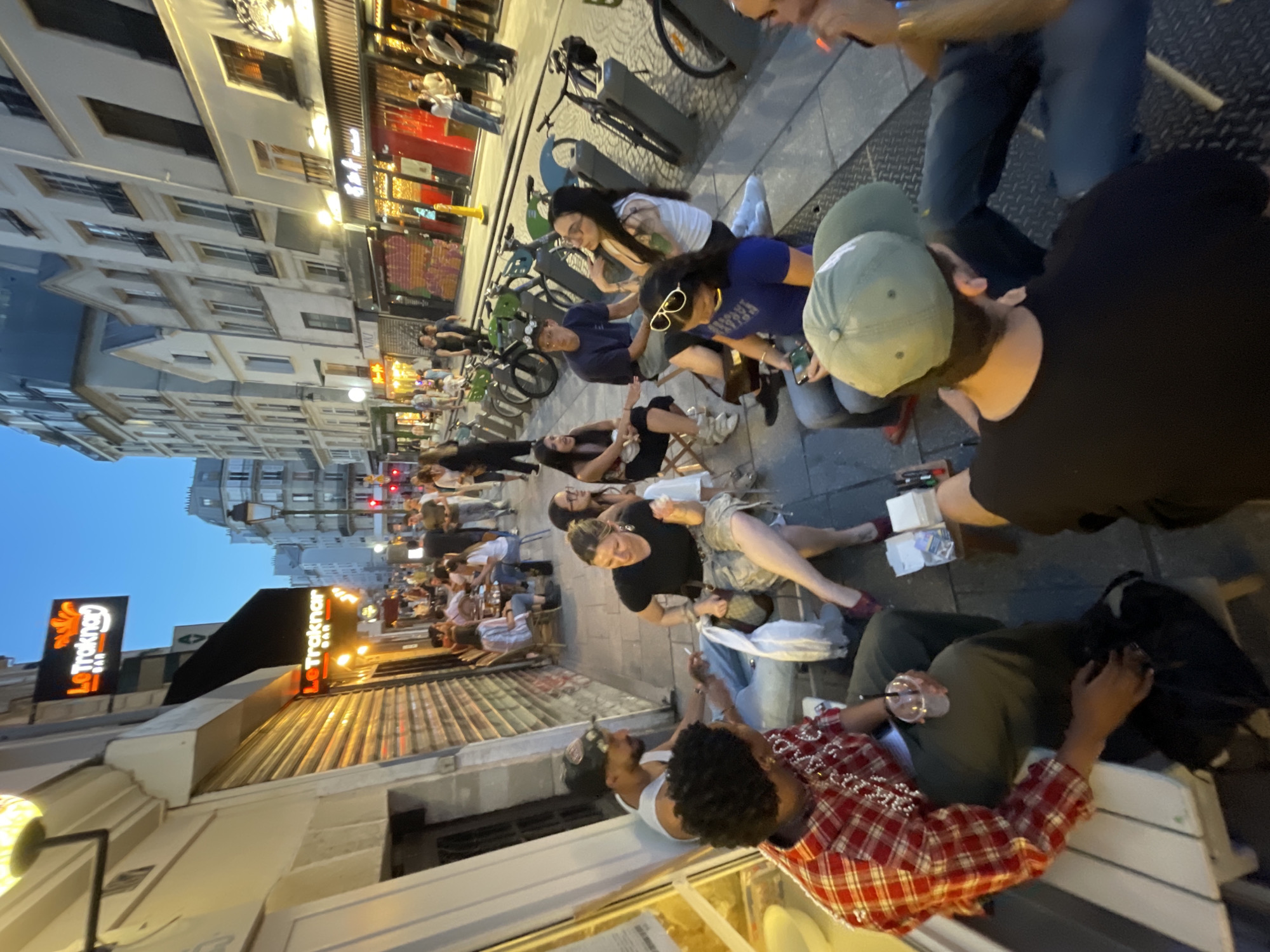 The image depicts an outdoor dining scene in Paris, France. Patrons are seated at tables along the sidewalk in front of "Le Traknar" bar. The scene extends down a narrow street lined with buildings. People are sitting, talking, and enjoying drinks. Bicycles are parked nearby. The sky appears to be in the early evening twilight.