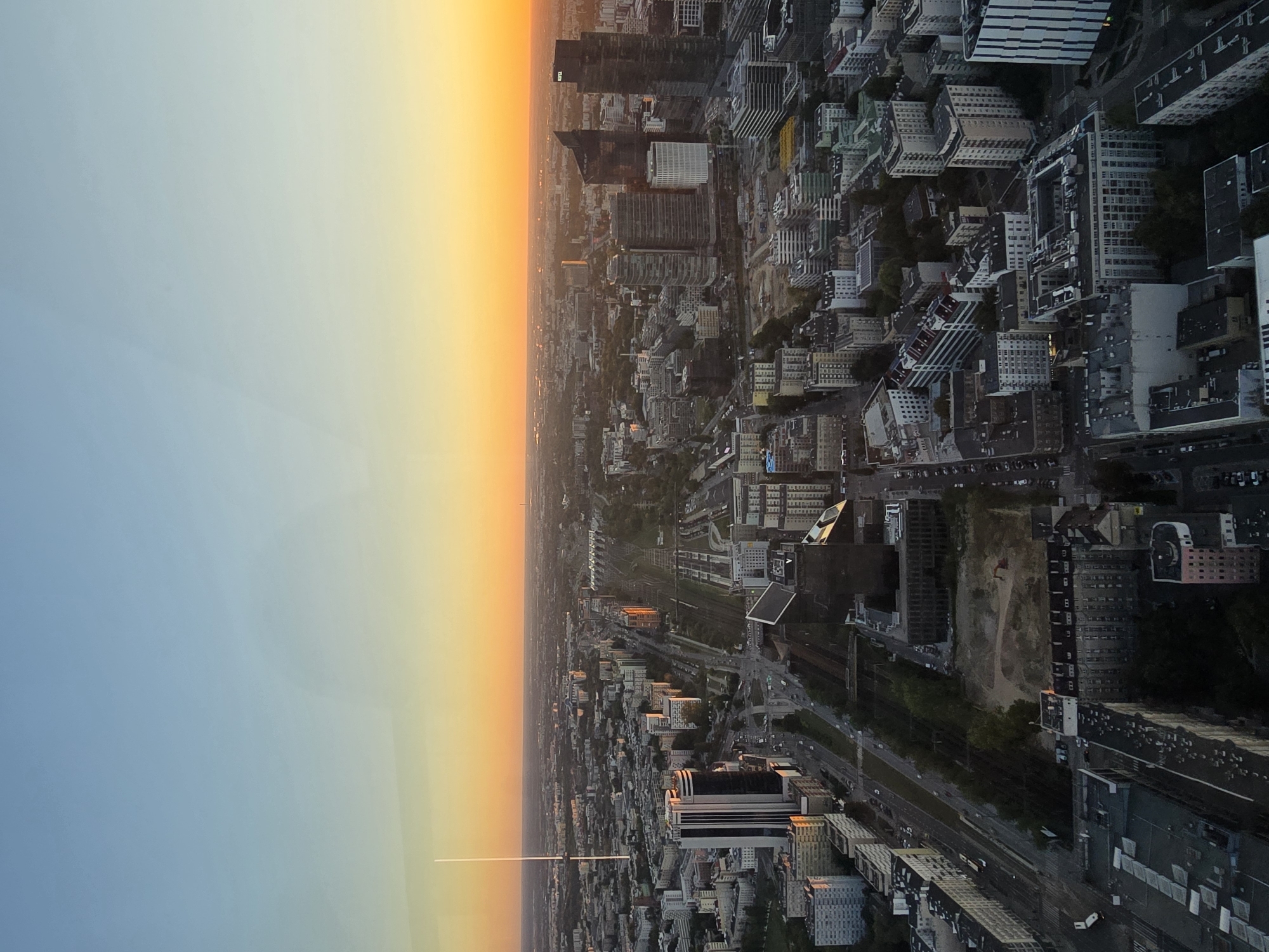 An aerial, high-angle shot captures the cityscape of Warsaw, Poland, during a sunset. The sky is a gradient of soft blue at the top, transitioning to a vibrant orange and yellow near the horizon, casting a warm glow over the urban landscape. The foreground is dominated by a dense collection of buildings, ranging from multi-story residential blocks to modern skyscrapers.
A prominent feature is a long, slender radio tower with a light at its apex, standing tall against the sunset. The city is crisscrossed by roads with visible traffic, and a railway line with multiple tracks runs through the scene, disappearing into the distance. Some buildings show reflections of the sunset, adding to the visual appeal. The overall impression is of a sprawling metropolis at dusk, bathed in the soft light of a setting sun.