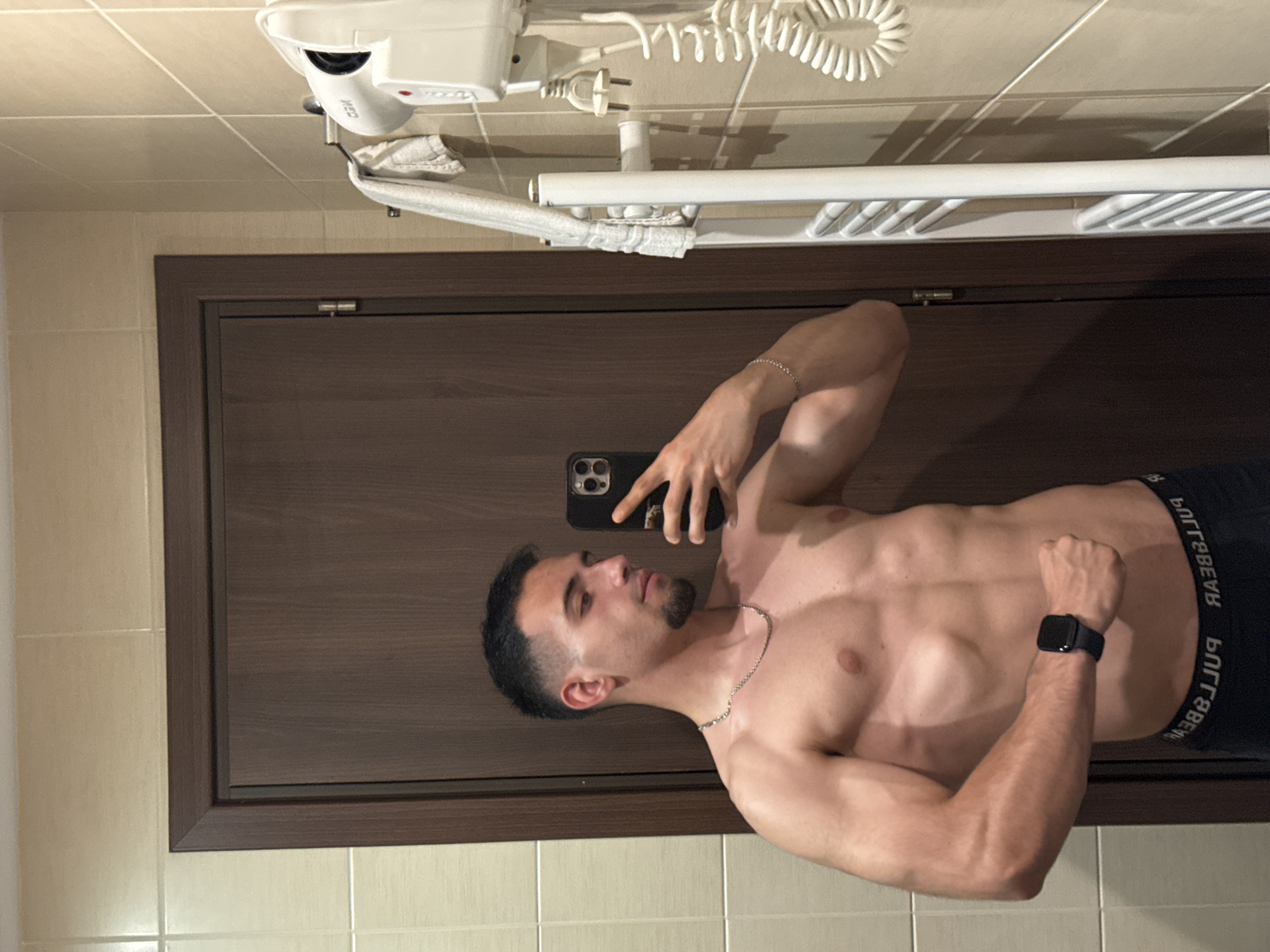 A shirtless man is taking a selfie in a bathroom. He is wearing black underwear and a watch. The bathroom has a wooden door, white tiles, and a white towel rack with a hair dryer. There is no information to indicate the location is in Veliko Tarnovo, Bulgaria.