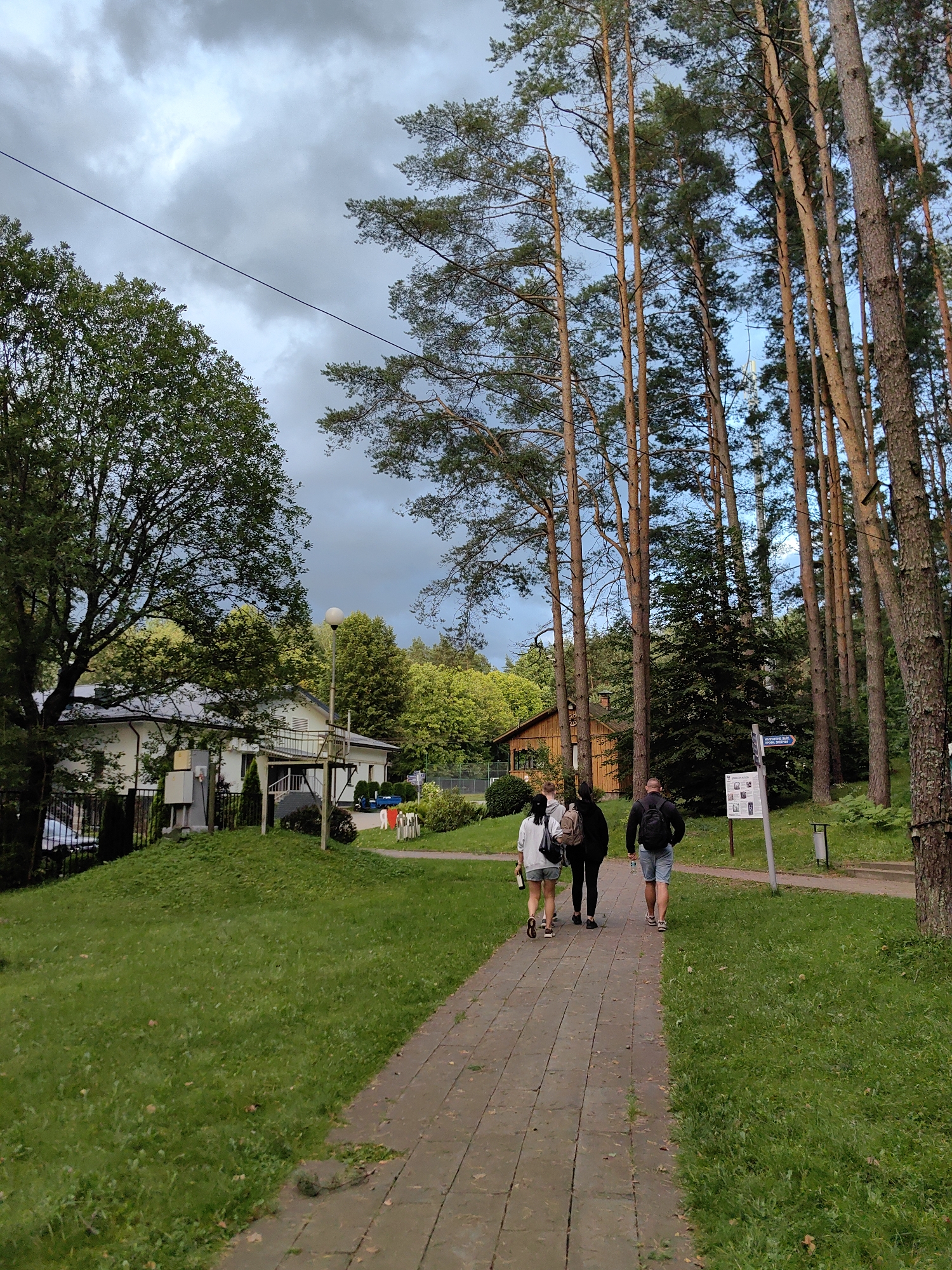 The image depicts a paved pathway in a park-like setting, bordered by a lawn on the left and tall pine trees on the right. Four individuals are walking away from the camera on the pathway, heading towards a signpost. The person in the front is wearing a black backpack and light blue shorts. To their left are two individuals, one with a backpack and the other without, both wearing light-colored clothing. The fourth person, furthest to the left, is wearing a light-colored jacket and shorts. In the background, a white building with a dark roof and a raised walkway is visible amongst trees. Further in the distance, a wooden structure with a brown roof is nestled among the trees. The sky is overcast with a mix of grey and lighter clouds. The scene appears to be in the vicinity of the Ракаўскі сельскі Савет in Belarus.