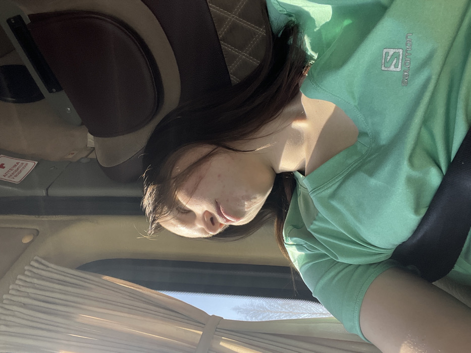 A young adult female is depicted in profile, seated inside a passenger vehicle, likely a bus or large van, in Kazanskii Kliuch, Russia. She has dark hair and visible skin blemishes on her right cheek and jawline. Her eyes are closed, and her head is tilted back and to her left, suggesting she is asleep or resting. She is wearing a light teal short-sleeved V-neck shirt with a white "Salomon" logo and text on the upper right chest. A black vehicle seatbelt is fastened diagonally across her chest.
Strong natural sunlight streams into the vehicle from a window on the left, illuminating the left side of her face, neck, and shoulder, and creating distinct shadows. The window is partially obscured by light-colored, pleated curtains tied back. Through the window, a clear blue sky is visible, with faint outlines of what appear to be bare trees or branches in the distance. To the right, behind the female, an upholstered seat with brown and lighter beige sections, featuring a quilted pattern, is visible. Above the window, a white and red sign with a medical cross and Cyrillic text, reading "АПТЕЧКА НАХОДИТСЯ У ВОДИТЕЛЯ" (First Aid Kit Located with the Driver), is affixed to the interior panel. The overall lighting suggests daytime.