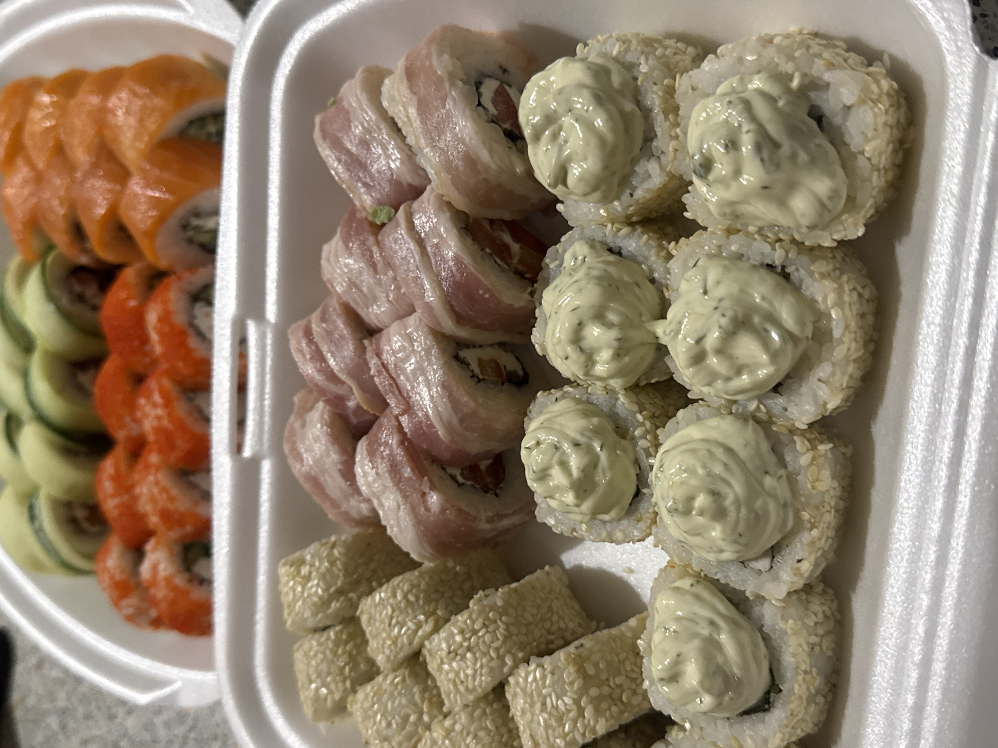 The image shows a close-up, overhead view of a white styrofoam container filled with various types of sushi rolls. The container is divided into two sections. The top section contains several rows of sushi rolls, some topped with orange roe, others with what appears to be cucumber and possibly avocado. The bottom section of the container holds different sushi rolls. One group of rolls is covered in a creamy green sauce. Another group of rolls is wrapped in thin slices of raw pink meat, possibly bacon or ham. The background is blurred, but appears to be a countertop. There is no discernible indication of the city of Zaokskiy, Russia within the image.