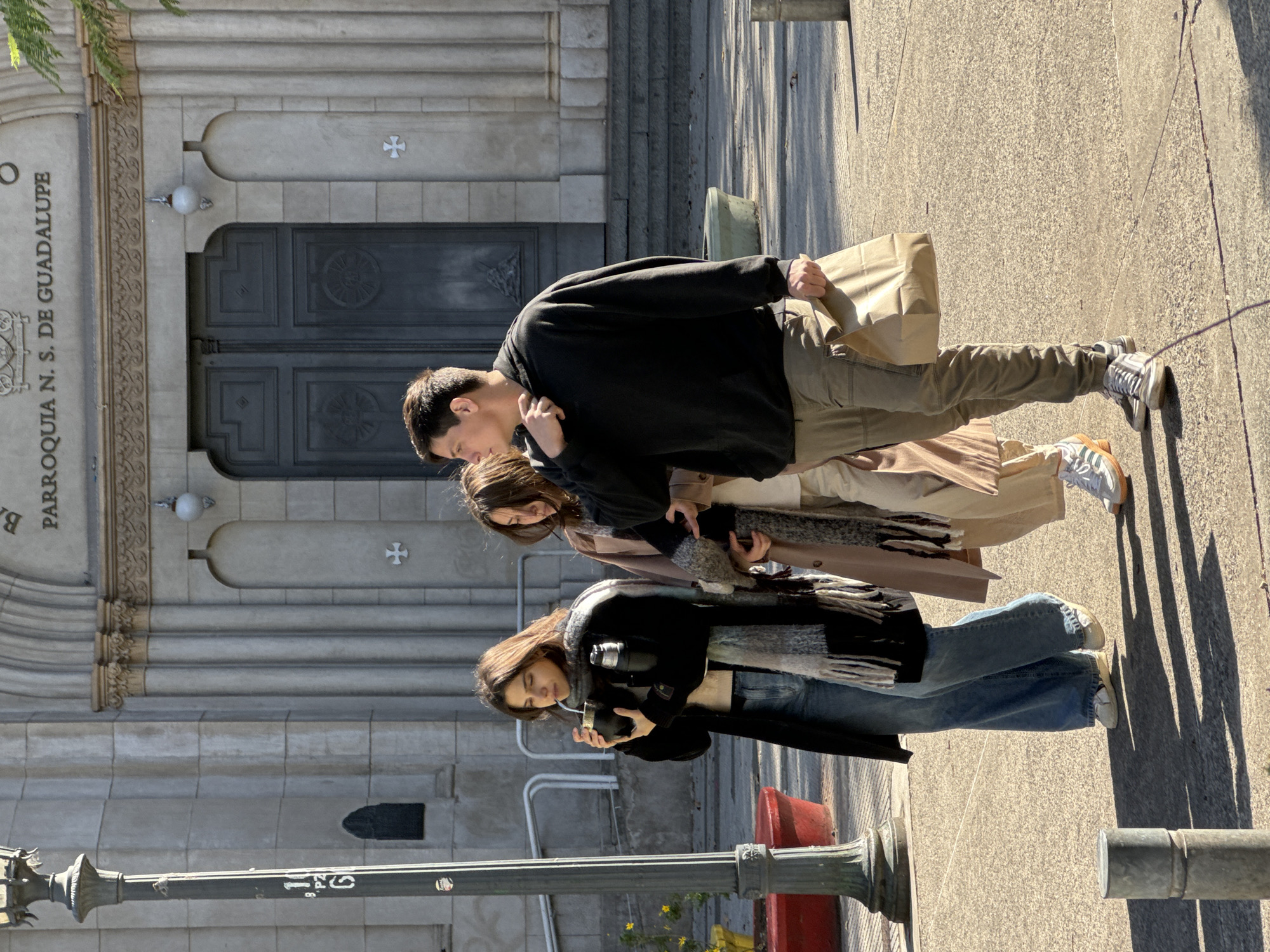 Three people stand on a sidewalk in front of a building with the inscription "PARROQUIA N. S. DE GUADALUPE." The person in the front has long brown hair and is looking down while holding an object. The person in the middle has long brown hair and is also looking down. The person in the back, a male, has short dark hair and is holding a tan paper bag. The setting is most likely Buenos Aires, Argentina, due to the inscription on the building.