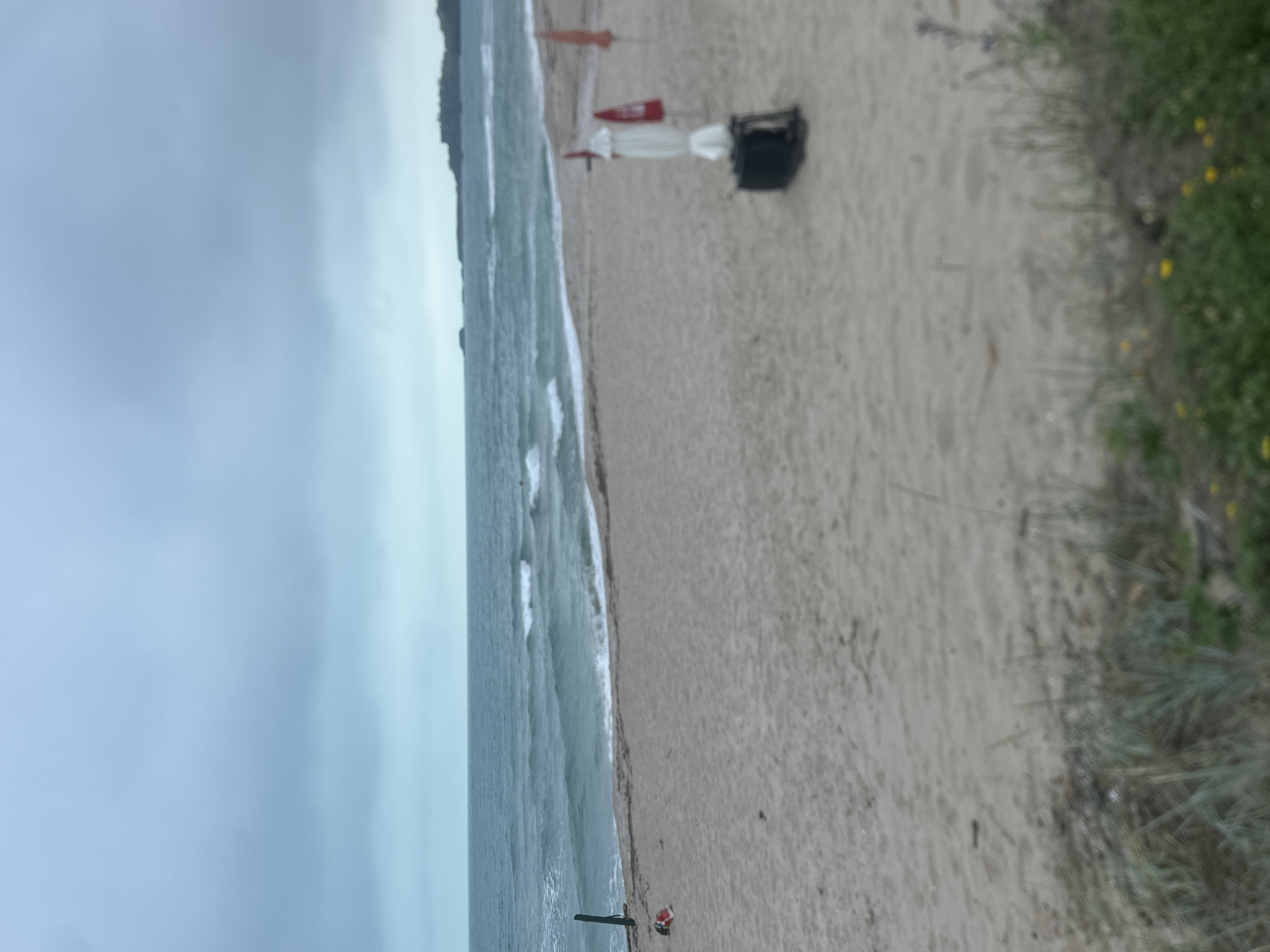 The image depicts a serene beach scene on an overcast day, captured from a slightly elevated vantage point looking down the coastline.
The sky, occupying the top portion of the frame, is a uniform light grey, indicating heavy cloud cover. To the left, the sea stretches horizontally, displaying muted blue-green waters with gentle waves breaking onto the sandy shore, creating lines of white foam. A very small, indistinct figure might be visible in the water in the lower left section.
The central and largest part of the image is the expansive sandy beach, light beige in color, extending from the water's edge towards the right. The sand shows subtle ripples and faint imprints.
Various objects are sparsely distributed on the sand. Towards the middle-right, further up from the water, there are two red, triangular shapes, resembling small flags or marker buoys, positioned upright. Nearby, a closed white beach umbrella stands next to a dark, rectangular object that could be a storage box or a covered shelter. Closer to the bottom-center of the image, near the waterline, a dark, elongated object, possibly a paddleboard or a small boat, rests on the sand next to a small red item, perhaps a cooler. A tiny, indistinct person appears to be seated on a low chair beside these objects, facing the water.
Along the far right edge of the frame, a strip of green vegetation, likely beach grasses or small dunes, runs parallel to the beach. Some small yellow flowers are visible among the grass.
The overall impression is one of a quiet, uncrowded beach, perhaps on a cooler or off-season day, given the overcast sky and minimal activity. There is no visible text in the image.