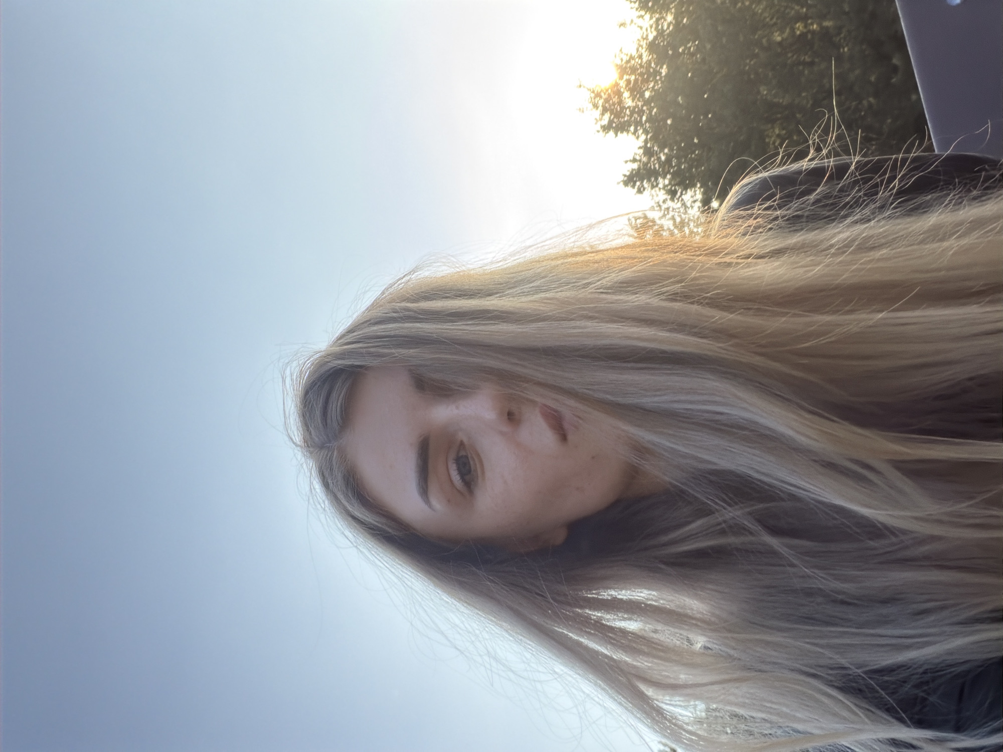 An outdoor, close-up, low-angle, selfie-style portrait shows a young woman with long, blonde hair that is blowing in the wind. Her hair obscures part of her face, but her left eye is visible, along with part of her nose and mouth. Her skin appears to have freckles. The background is a clear, light blue sky, with a hint of sun flare visible in the upper right corner, behind a cluster of green trees. The woman is wearing dark clothing.