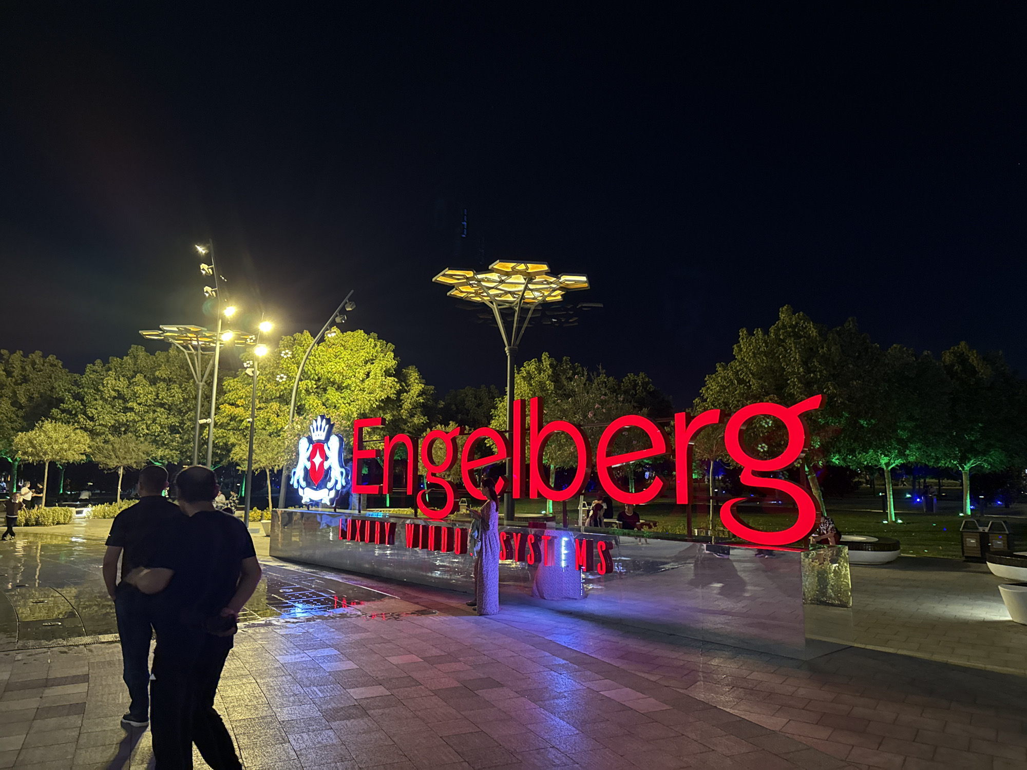 The image depicts a nighttime scene in Tashkent, Uzbekistan, specifically in the Tashkent City park. In the foreground, two men walk arm in arm, facing away from the camera, on a wet, tiled pathway illuminated by red and blue lights. Behind them, a large, illuminated red sign spells out "Engelberg." To the left of the sign, a crest-like emblem is also lit up. The background features trees with green uplighting, modern streetlights with unique hexagonal designs, and several people scattered throughout the park. The ground appears to be a mix of tiles and reflective surfaces, possibly water features. The overall atmosphere is festive and modern, highlighting a popular urban gathering space.
