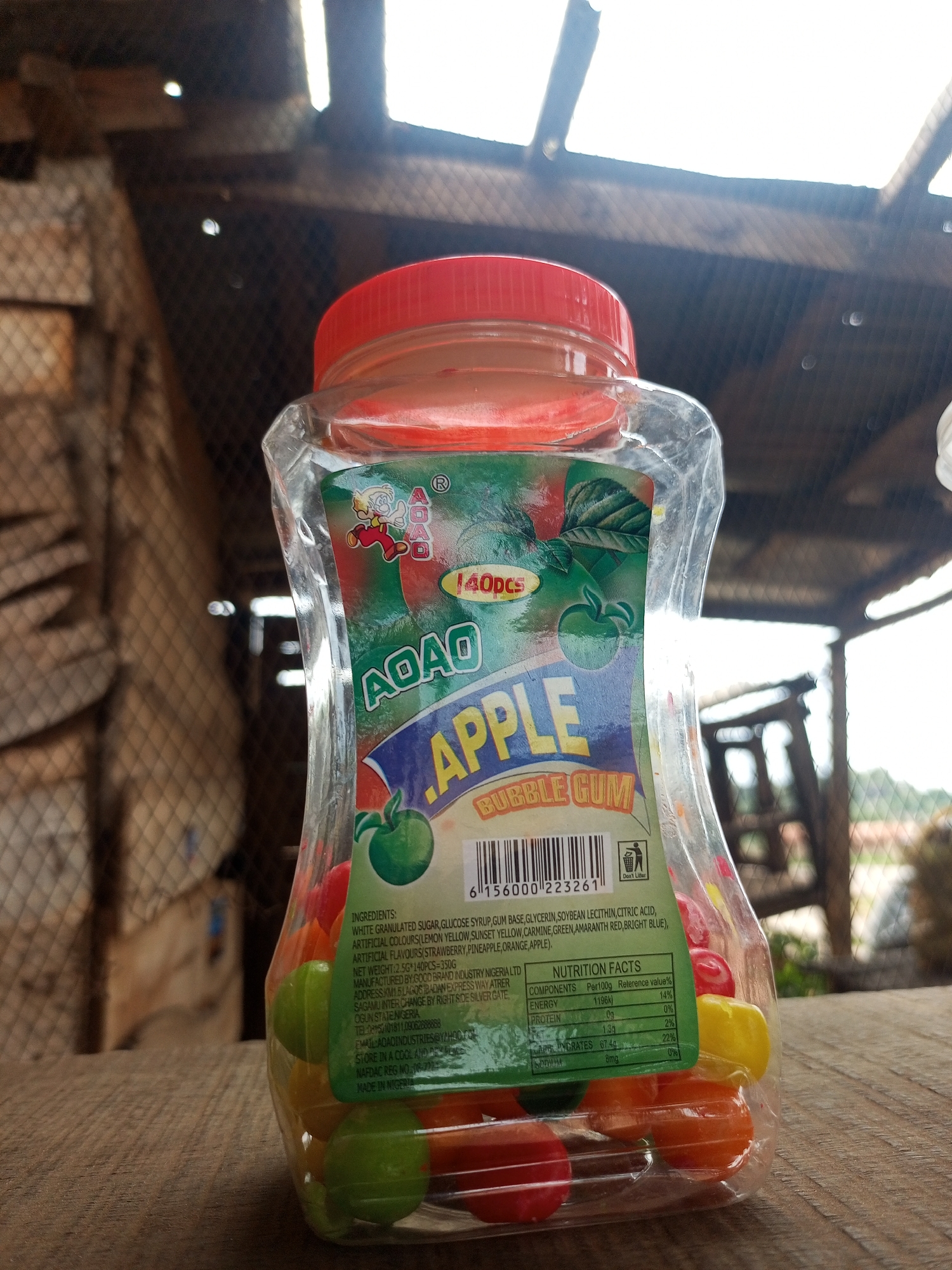An eye-level, medium outdoor shot shows a clear plastic bottle of "AOAO Apple Bubble Gum" standing on a wooden surface. The bottle is filled with colorful, spherical pieces of bubble gum in red, orange, green, and yellow. A red lid seals the bottle. The label on the bottle features cartoon characters and text detailing ingredients, net weight, manufacturer, and nutrition facts. Behind the bottle, a wire mesh fence is visible, with wooden structures and some greenery in the blurred background. The lighting suggests it is daytime, with light filtering from above and behind.