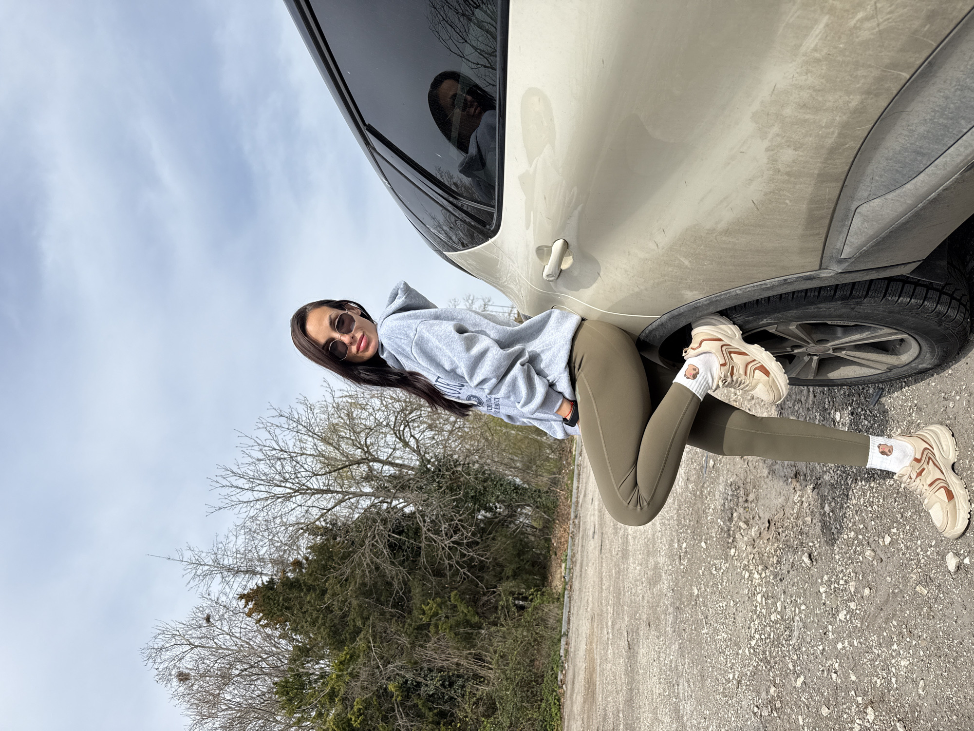 A young woman in sunglasses, a grey hoodie, and olive green leggings leans against a tan-colored SUV on a gravelly surface. She wears bulky off-white sneakers with brown accents and white socks featuring cartoon-like images. Trees with bare branches are visible in the background under a cloudy sky. The photo's angle suggests it was taken from a low vantage point and subsequently rotated. There is no specific information available in the image to confirm this location is Pleven, Bulgaria.