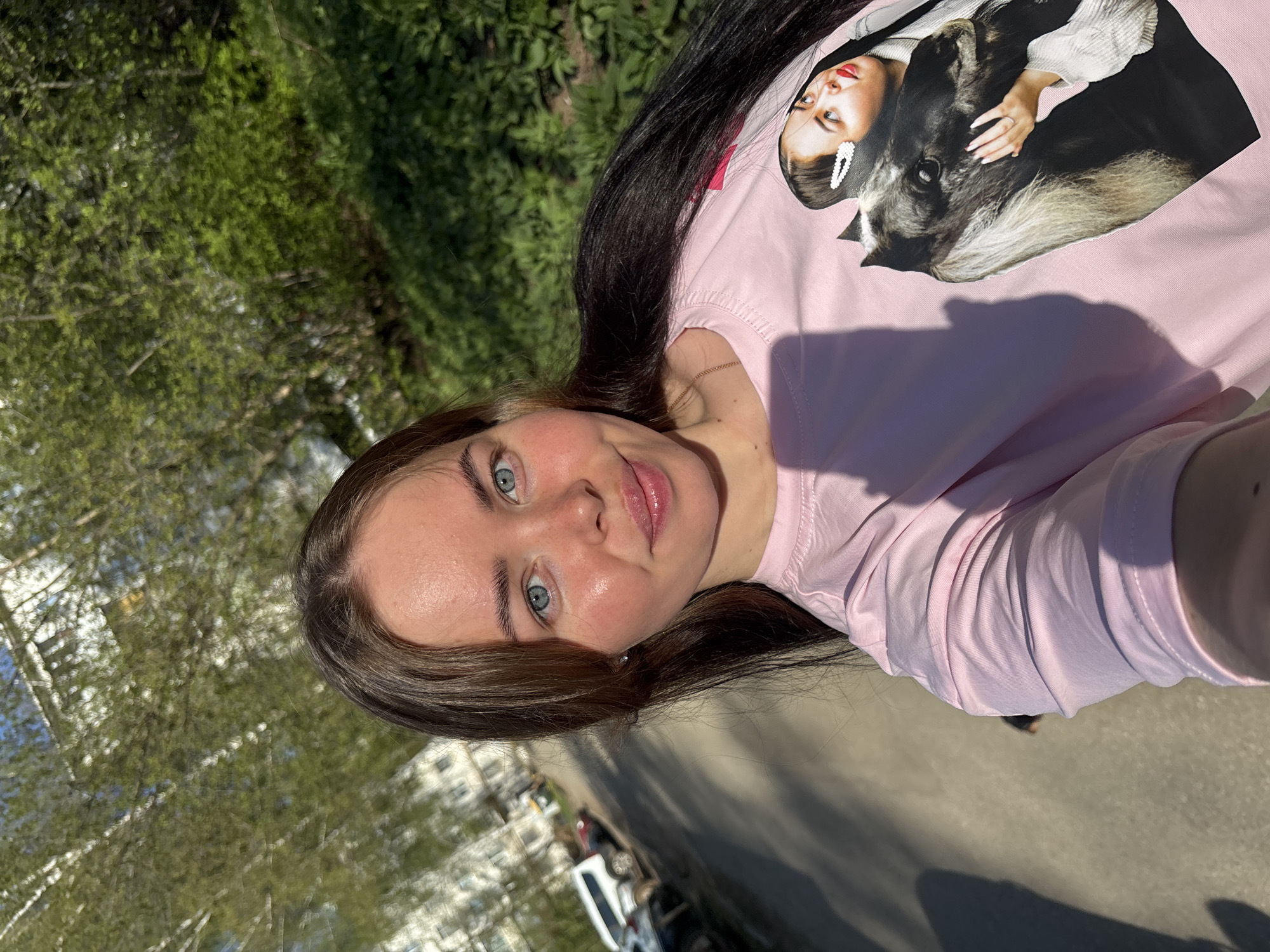 A young woman is outdoors, apparently standing on a sidewalk in Izhevsk, Russia. She is wearing a pink long-sleeve shirt with a graphic of a woman holding a dog. Her hair is dark and straight. The environment includes green trees and buildings in the background. The sidewalk is sunlit, casting shadows.