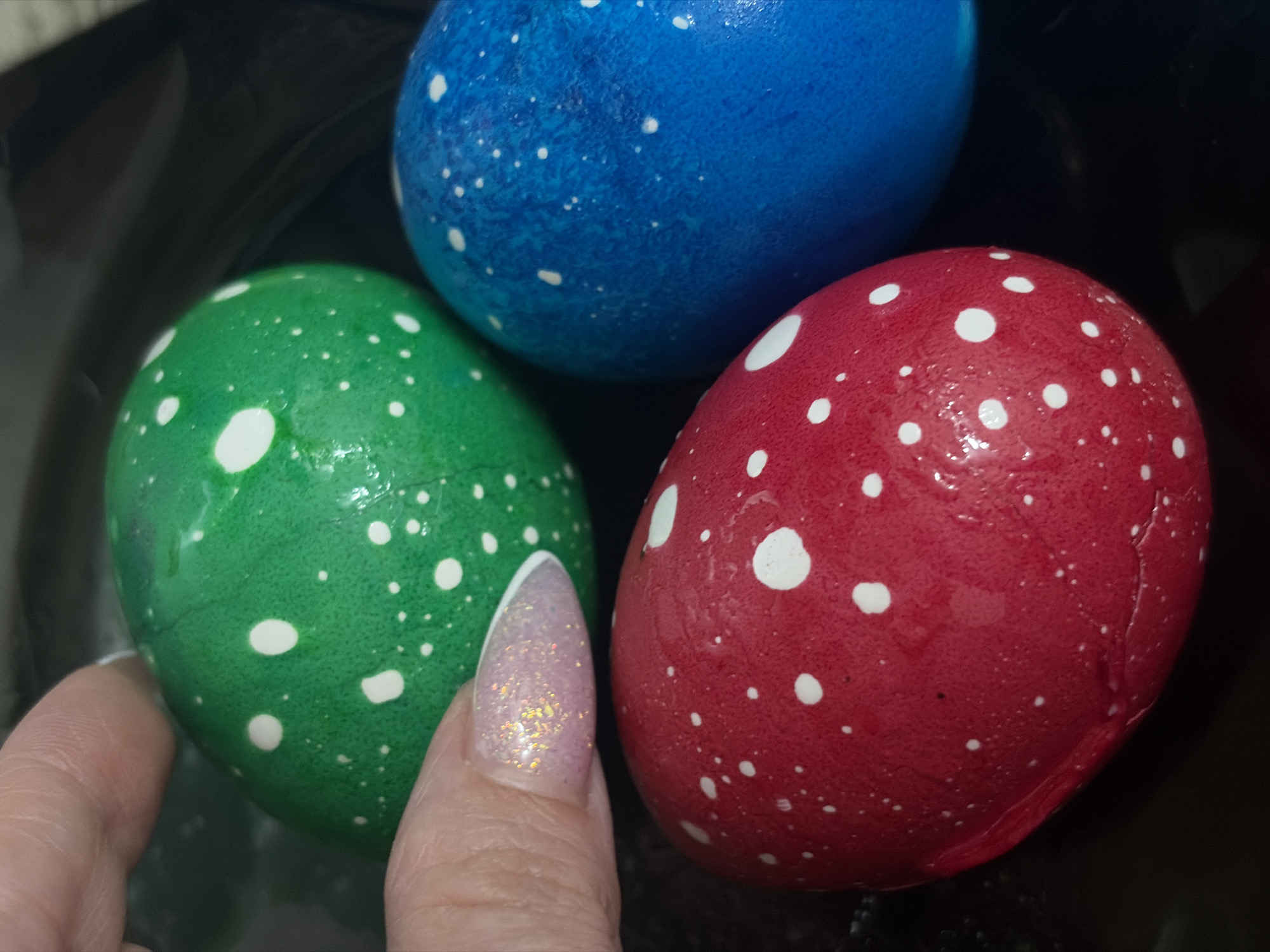 A close-up shot displays three Easter eggs, colored green, red, and blue, each speckled with white dots. A finger with a glittery pink and white nail is holding the green egg. The eggs are shiny, suggesting they may be wet or varnished. The surface they are placed on appears to be a dark, possibly black, color. There is no information to conclude anything about the location of Saint Petersburg, Russia.