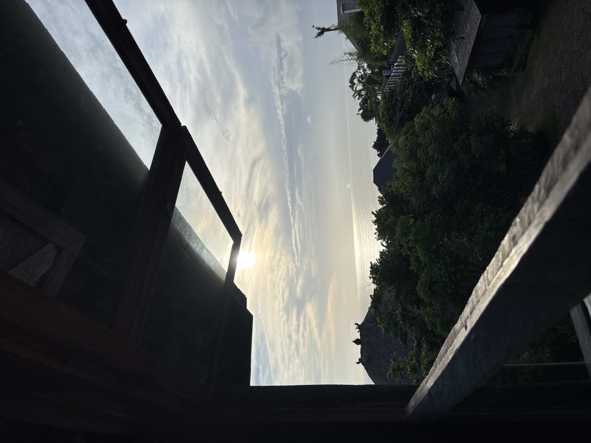 The image depicts a view from within a dark, wooden structure, possibly a balcony or an open-air building. The framing of the structure encloses a scene of the ocean, illuminated by the sun near the horizon, and the sky with light clouds. To the right of the ocean view, there is lush green vegetation and glimpses of rooftops, suggesting a tropical setting. The location is Kuta Selatan, Indonesia, indicated by the style of the architecture and the tropical flora. There are no people visible in the image. The primary subjects are the ocean, sky, and the vegetation, viewed through the wooden frame.