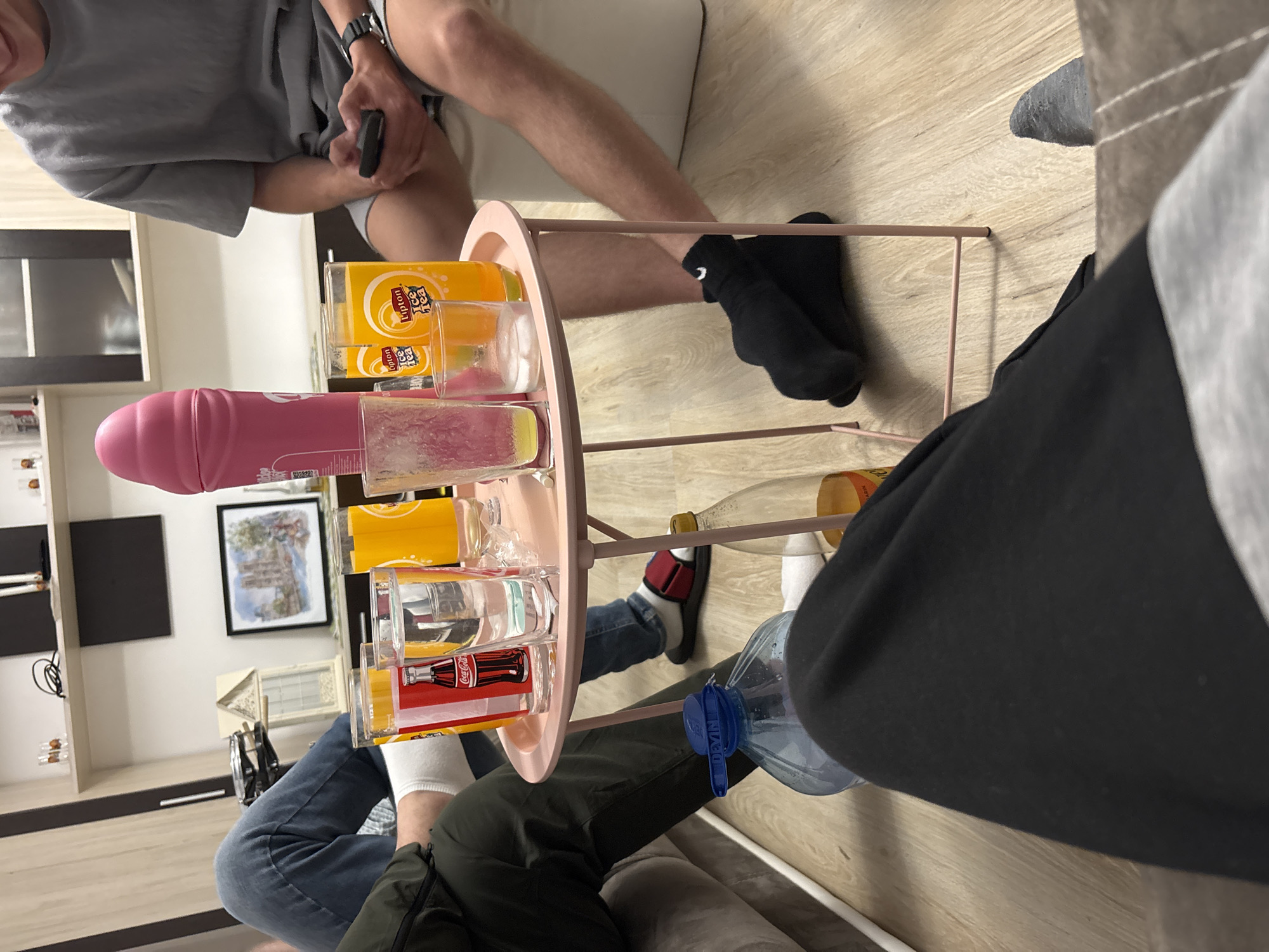The image shows an indoor scene, with several people seated around a small, pink, two-tiered serving cart filled with various glasses and bottles.
People: The image captures portions of at least three individuals. One person wears a grey t-shirt and shorts, holding a black object in their hand. Another is wearing black sweatpants. A third person is wearing jeans and an olive green jacket.
Objects: A pink serving cart holds several items, including a pink dildo, glasses with the logos of Coca-Cola and Lipton Ice Tea, and what appears to be a large bottle of water. A framed picture hangs on the wall.
Environment: The scene appears to be inside a home, with visible furniture and décor. A wooden floor is visible, and a cabinet is positioned against the wall.
The location in the city of Graf Ignatievo, Bulgaria is unknown.