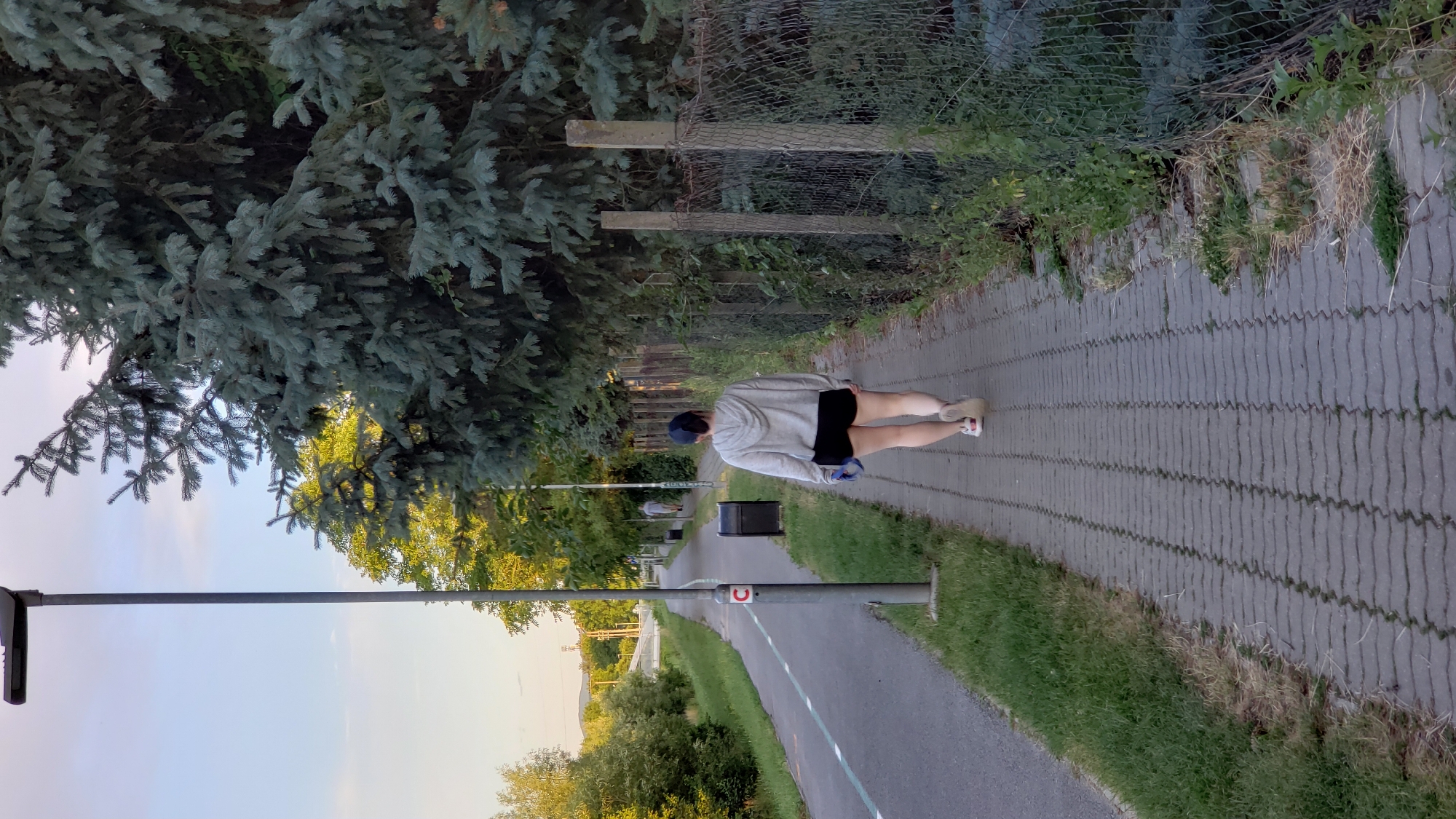 A person walks on a brick sidewalk next to a paved path in Prešov, Slovakia. The person is viewed from behind and wears a gray hooded sweatshirt, black shorts, a blue baseball cap, and white sneakers. They are carrying a blue object in their left hand. A street lamp is to the left of the person, and a wire mesh fence with concrete posts is to the right. There are trees and grass on both sides of the path and sidewalk. The sky is clear with some clouds. The scene appears to be a recreational area or park within the city.