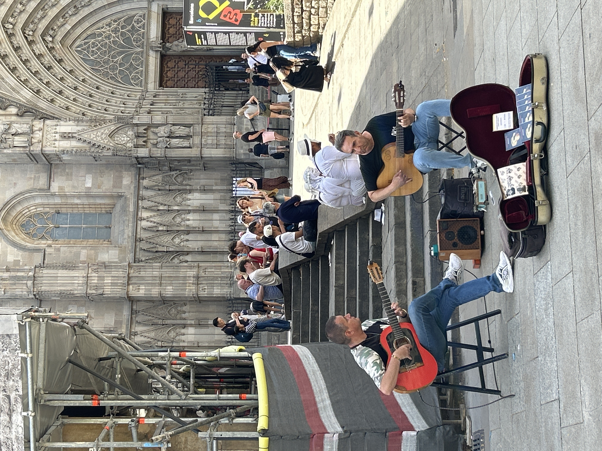 The image depicts a street scene in Barcelona, Spain, likely in front of a grand cathedral, given the Gothic architectural style of the building in the background. Two men are actively playing guitars in the foreground, positioned on the paved ground. The man on the left, seated on a black stool, wears a camouflage-patterned t-shirt and blue jeans, playing a red acoustic guitar. To his right, another man, also seated, plays a lighter-colored acoustic guitar and is dressed in a dark t-shirt and blue jeans. A partially open guitar case lies on the ground in front of him, containing several CDs and pamphlets.
A group of people are visible on stone steps behind the guitarists, some sitting and observing, others standing. Further back, more people are milling about, suggesting a public square or thoroughfare. Scaffolding is present on the left side of the image, indicating potential ongoing construction or renovation work on the building. The environment is sunny, casting shadows on the ground, and the paving stones are visible, suggesting an outdoor urban setting.