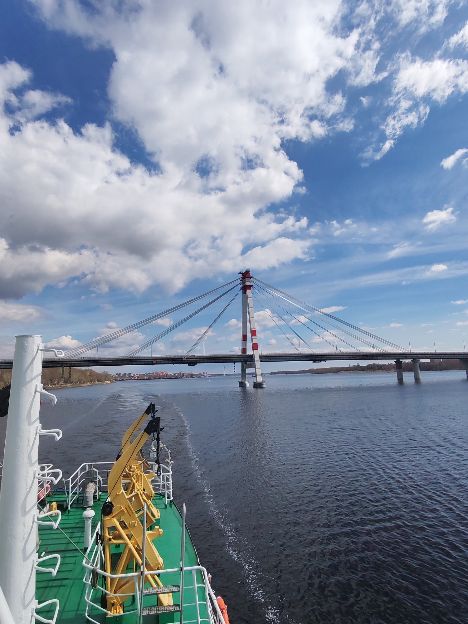 The image captures a view from a vessel on a body of water, with a bridge in the background. The bridge is a cable-stayed structure with a tall tower painted in red and white stripes. Cables extend from the tower to the bridge deck. Another section of the bridge extends to the right, supported by pillars in the water. The sky is partly cloudy with a mix of blue sky and white clouds. The water is dark with visible waves and the wake of the vessel. In the foreground, parts of the vessel are visible, including a green deck, white railings, and yellow machinery. The location is Riga, Latvia.