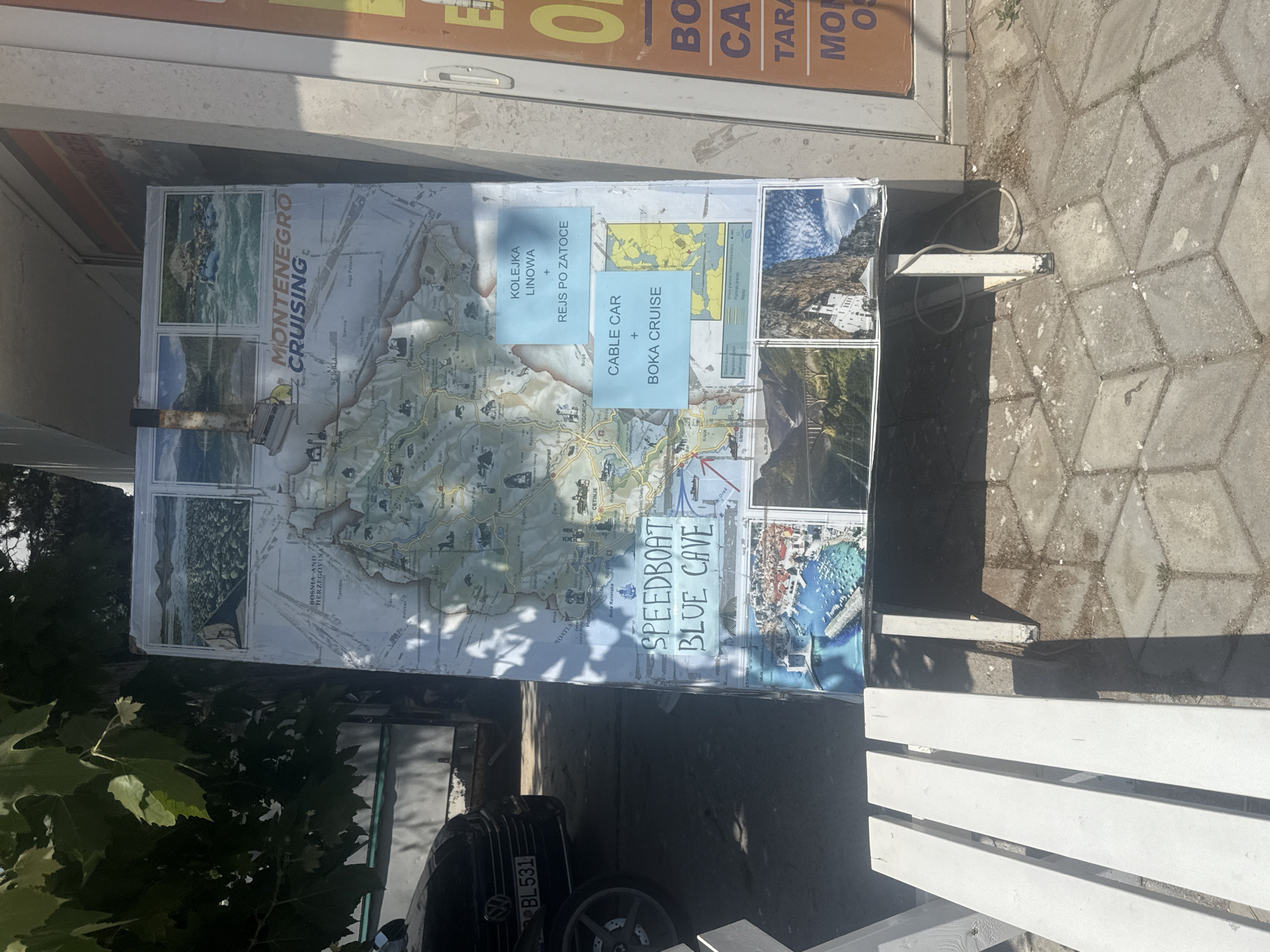 The image depicts an outdoor scene in Boreti, Montenegro, where a large, weathered informational board is displayed. The board, positioned on a paved area with a white bench in the foreground, features a map of Montenegro along with various photographs and text. Several smaller photos are arranged at the top and bottom of the board, showcasing landscapes and activities like white-water rafting and boat tours. Text on the board includes "MONTENEGRO CRUISING," "SPEEDBOAT BLUE CAVE," and information about a "CABLE CAR" and "BOKA CRUISE" in both English and what appears to be Polish ("KOLEJKA LINOWA + REJS PO ZATOCE"). The board itself appears to be standing on metal legs. To the left of the board, a black car with a license plate reading "BL 531" is partially visible. Trees with green leaves overhang the scene from above, casting shadows. The overall environment suggests a public space, possibly near a tourist attraction or information point.