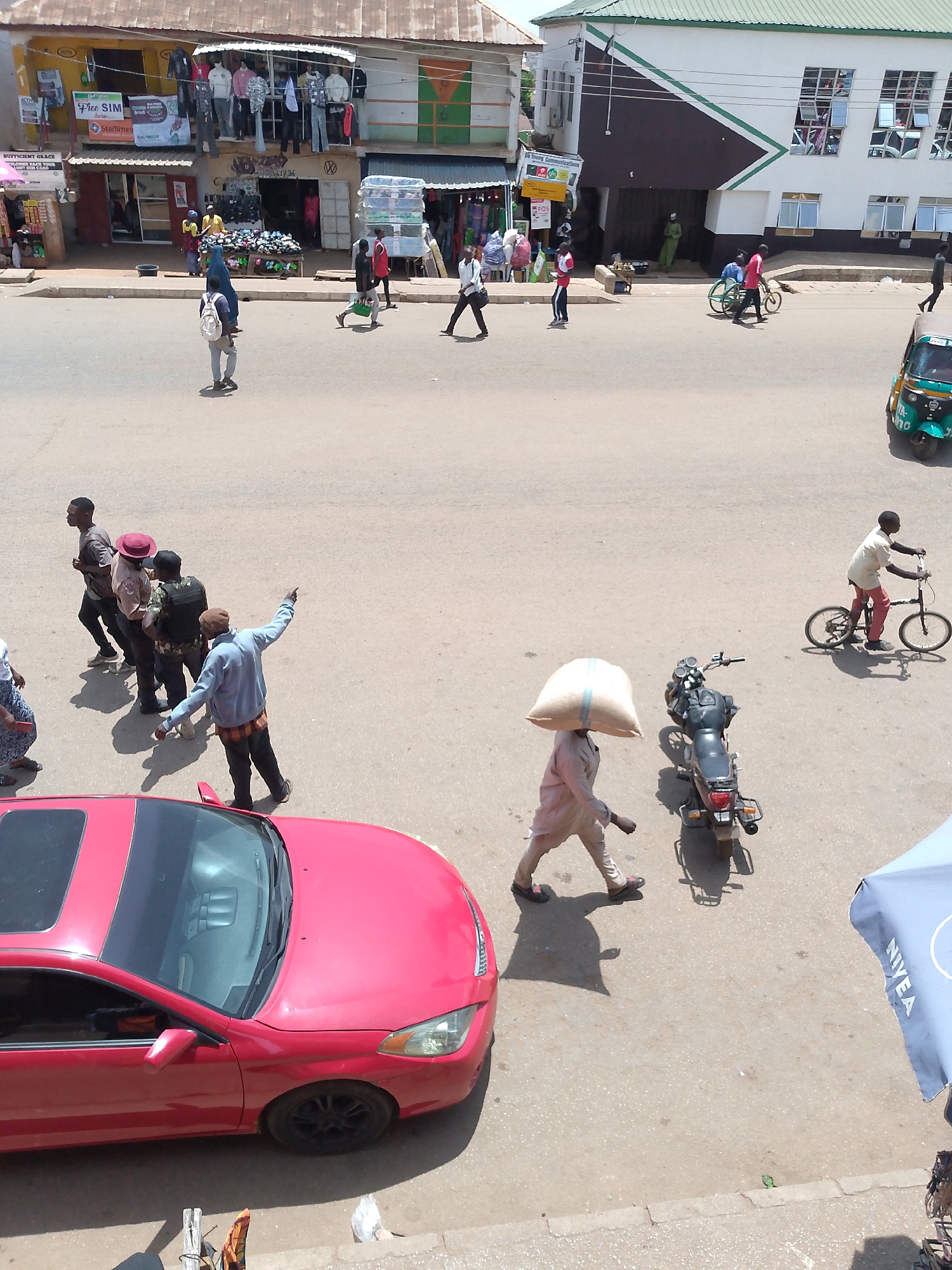 The image shows a street scene in Bukuru, Nigeria, taken from an elevated perspective. The street is wide and paved, with several people walking or cycling. Buildings line both sides of the street, including shops with awnings and displays of goods. A red car is parked on the left side of the frame. A man carries a large sack on his head near a parked motorcycle. Another person is riding a small bicycle. Other people are moving in various directions across the street. A tuk-tuk is visible on the right side of the image. There's a "Free SIM" banner visible on one of the shops. A Nivea-branded umbrella is present in the lower right corner.