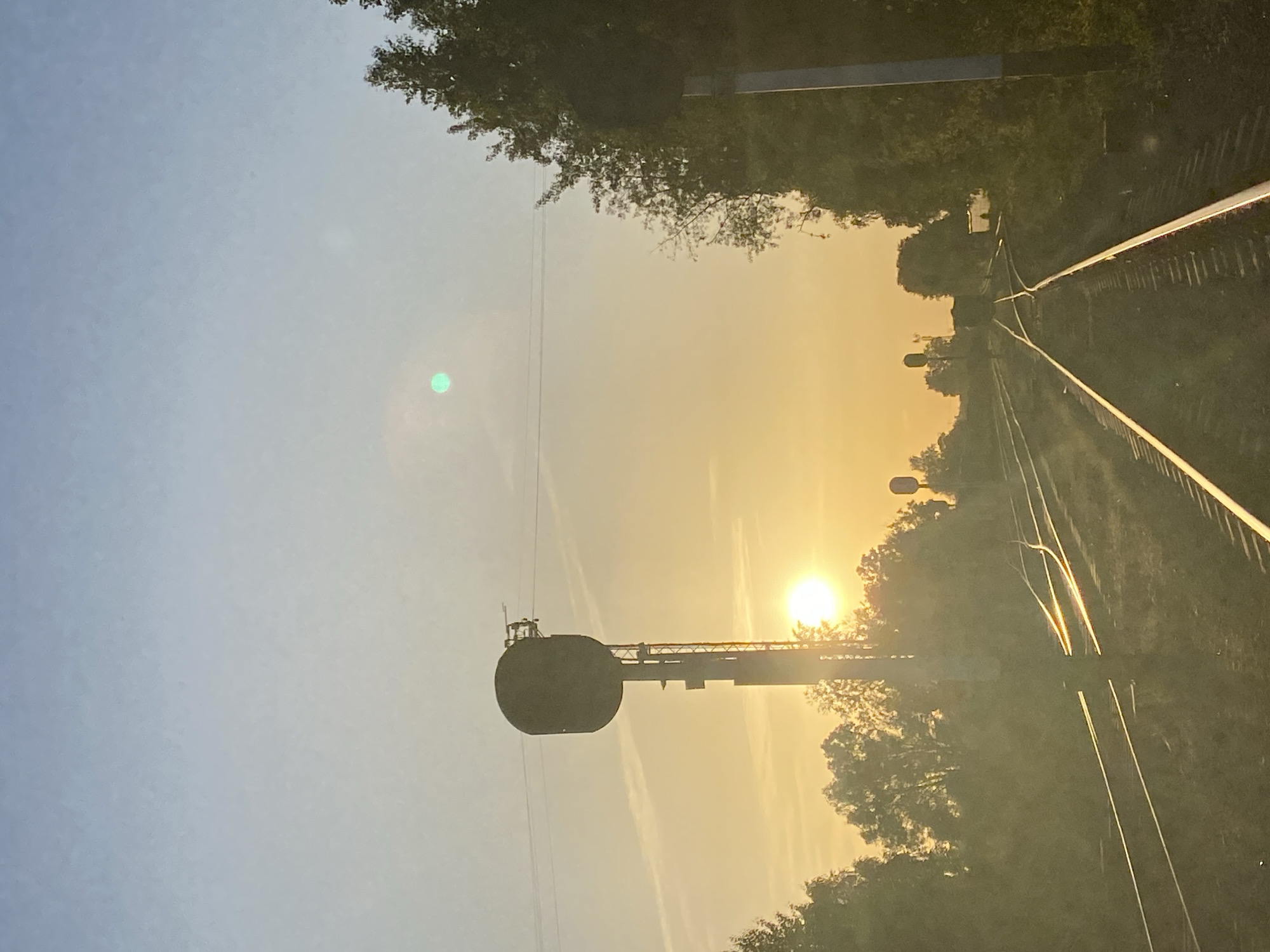 A railway runs through a tree-lined area, with the sun shining at the end of the tracks. An overhead wire system with a large circular component is visible to the left. The location is likely Chaikovsky, Russia, based on the prompt.
