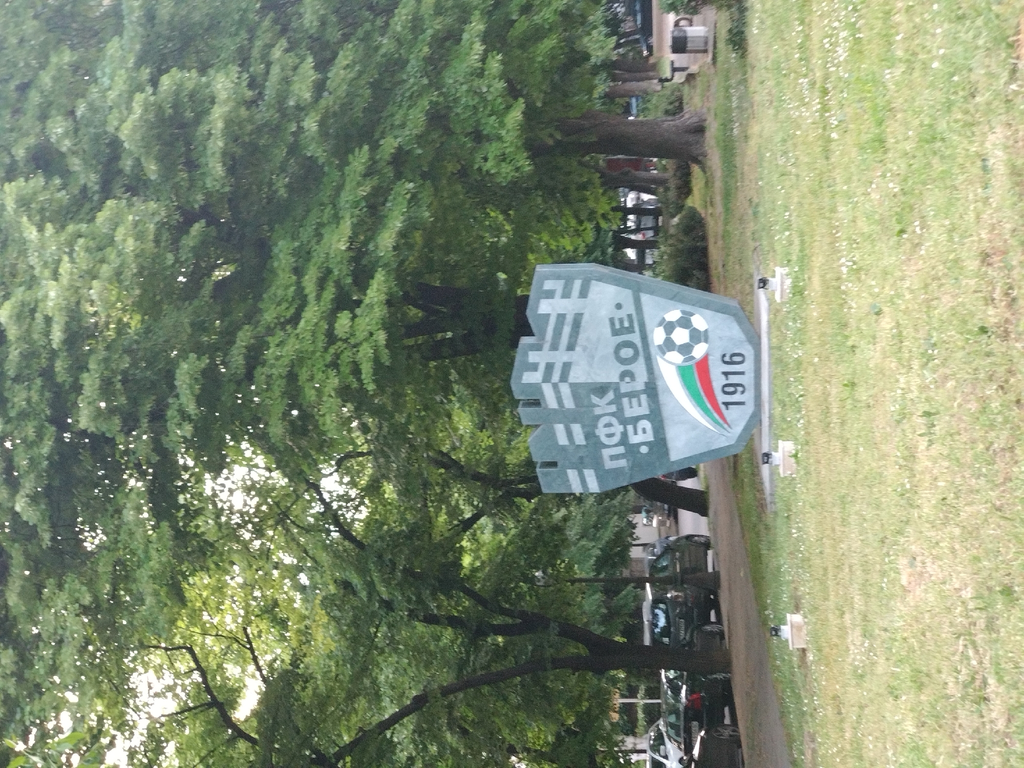 The image depicts a monument for the football club PFC Beroe Stara Zagora in Bulgaria. The monument, made of stone or concrete, is decorated with the club's logo, a football, and the year of its founding, 1916. It is located on a strip of grassy land next to a road with parked cars. Trees line the road and the grassy area. The monument is in the city of Stara Zagora, Bulgaria, based on the team associated with the monument.