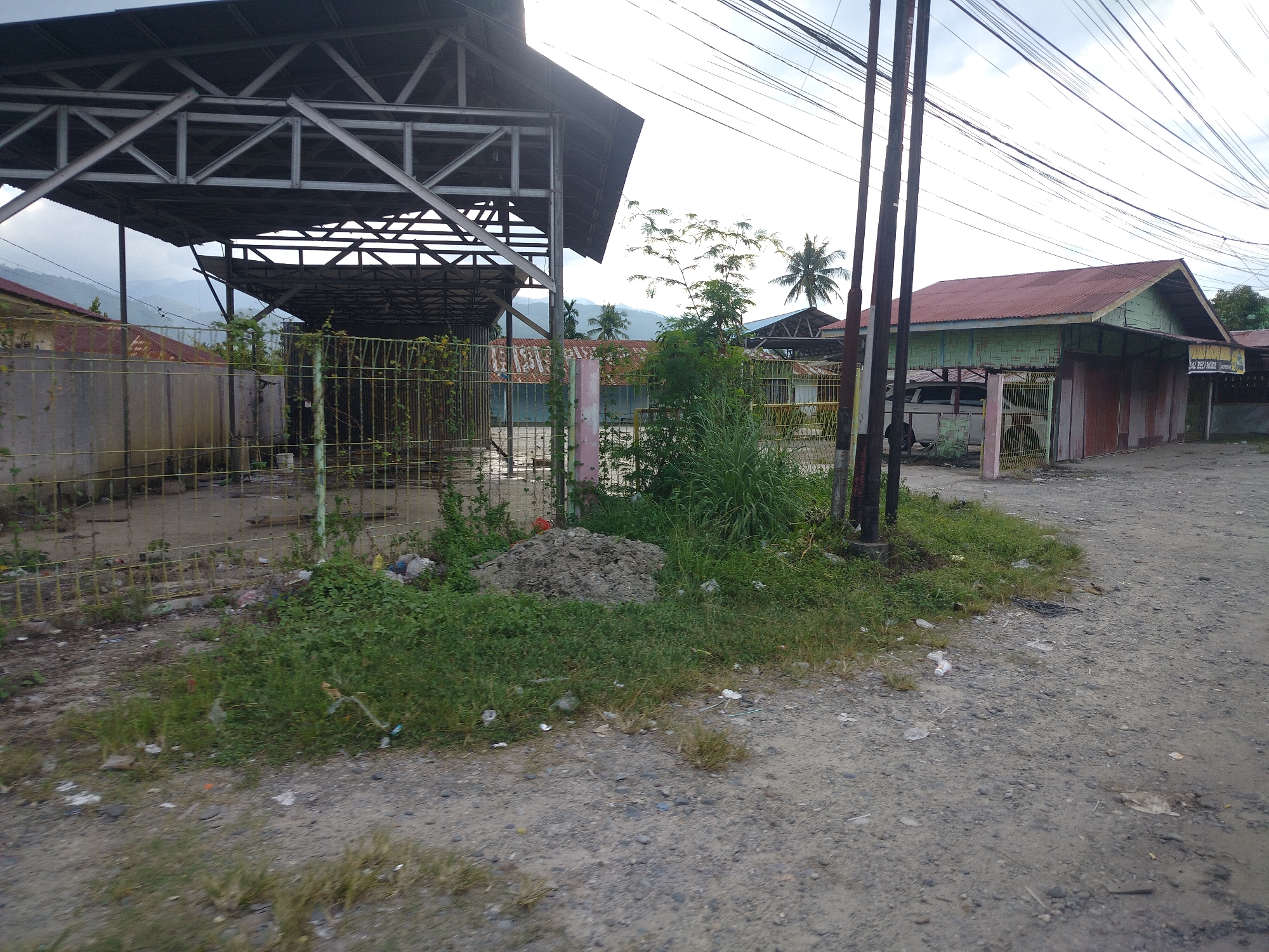 The image depicts an outdoor scene on a cloudy day in Kampungmelayu Satu, Indonesia. In the foreground, a dirt and gravel road is visible, with scattered debris including plastic bottles and wrappers. To the left, a yellow wire fence encloses an area with overgrown grass and a pile of dark soil. Beyond the fence, a large, open-sided structure with a metal frame and corrugated metal roof stands partially constructed or in disrepair. Several utility poles with a dense network of overhead wires are prominent on the right side of the image. In the middle ground, a low-slung building with a red corrugated metal roof and bamboo siding is visible, with its front partially obscured by a yellow gate. Further in the background, more buildings with similar roofs and a distant mountainous landscape under a hazy sky can be seen. The overall impression is one of a developing or somewhat neglected urban fringe environment.