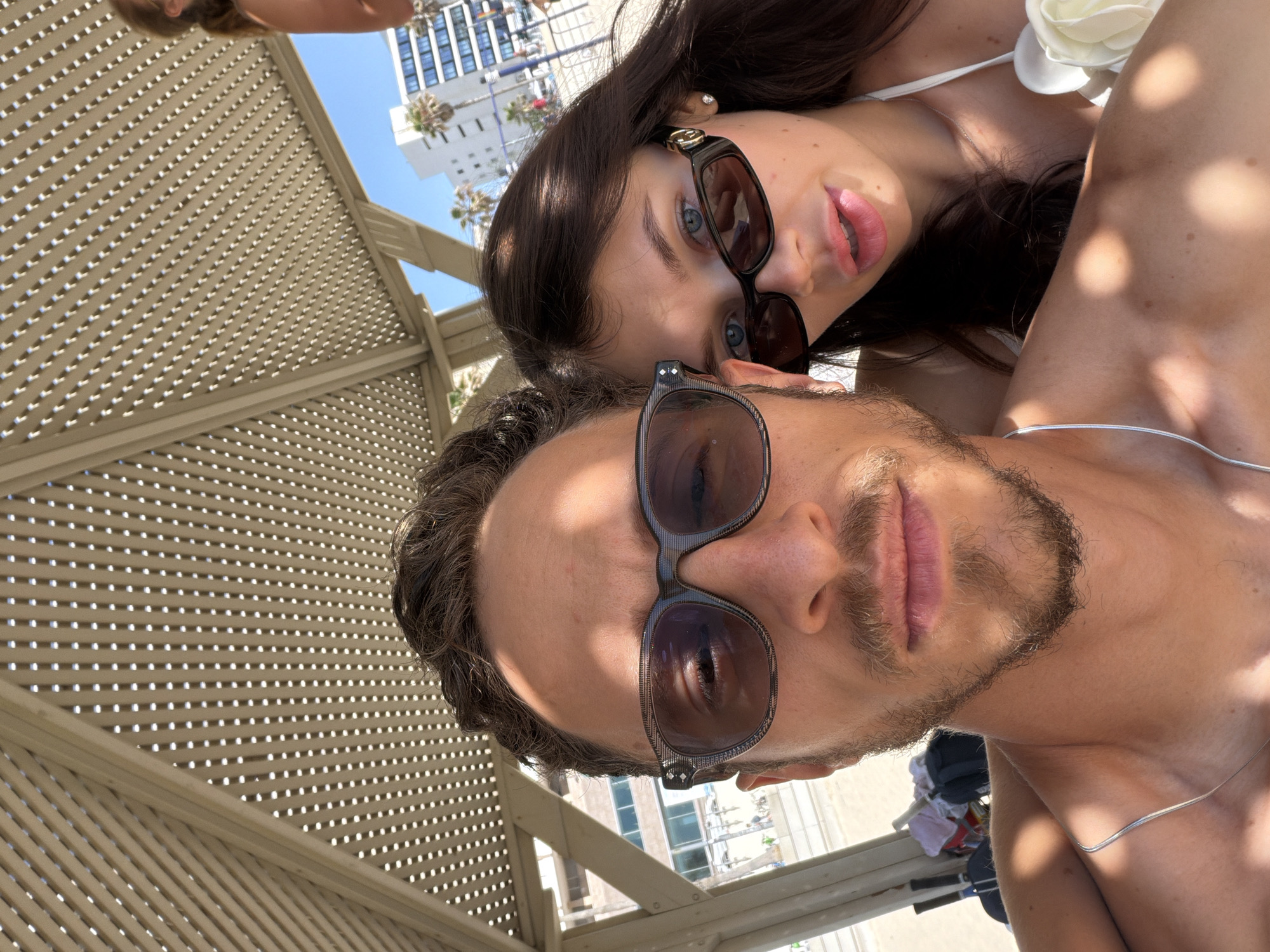 This is a close-up, selfie-style photograph depicting a male and a female individual under a slatted sunshade structure, located in Tel-Aviv, Israel, on a bright, sunny day.
The male, positioned in the foreground and center-left, has dark brown wavy hair, a short beard, and tanned skin. He wears dark-rimmed sunglasses, with a subtle pattern visible on the frames, and a thin silver chain necklace. His right eye is partially visible through his sunglasses, and he gazes directly towards the camera. His bare right shoulder and upper chest are visible, showing dappled sunlight and shadow patterns.
The female is positioned behind and to the right of the male. She has long dark brown hair, fair skin, and blue eyes. She wears dark, rectangular sunglasses featuring a gold-colored accent on the temples and a small stud earring in her right ear. Her lips are slightly parted. She is wearing a white bikini top adorned with a white floral appliqué on the right strap/cup area, and a thin silver chain necklace is partially visible on her chest. Her left shoulder and upper chest are visible.
The scene is illuminated by direct sunlight, filtered through a light-colored, slatted overhead structure, which casts distinct circular light patterns and elongated shadows on the subjects and surfaces. In the background, under a clear blue sky, several multi-story buildings are visible, including one with blue-tinted windows, indicative of modern urban architecture. Palm trees are also visible, consistent with a coastal city environment. Behind the male's left shoulder, a sandy area is visible, along with blurred objects that appear to be beach equipment.