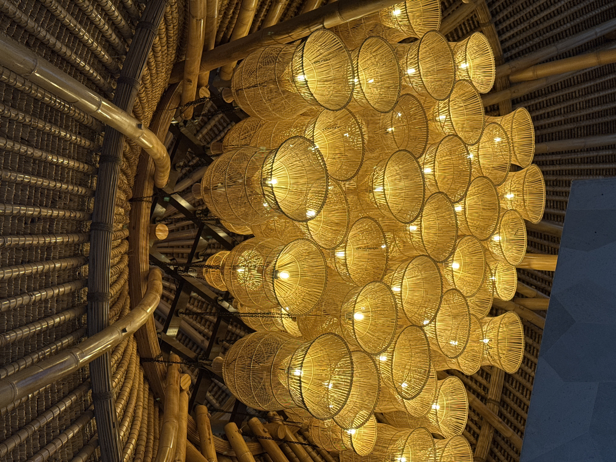 A high-angle interior shot depicts a large grouping of wicker lamps hanging from a bamboo ceiling in an unknown location in Indonesia. The wicker lamps are round with a cone-shaped bottom and are lit from within, casting a warm glow. The bamboo ceiling is constructed of rows of bamboo poles and woven bamboo sheets. The composition is asymmetrical, with the lamps concentrated in the center and right of the frame, while the bamboo ceiling fills the left side. A gray wall is visible on the right edge of the image.