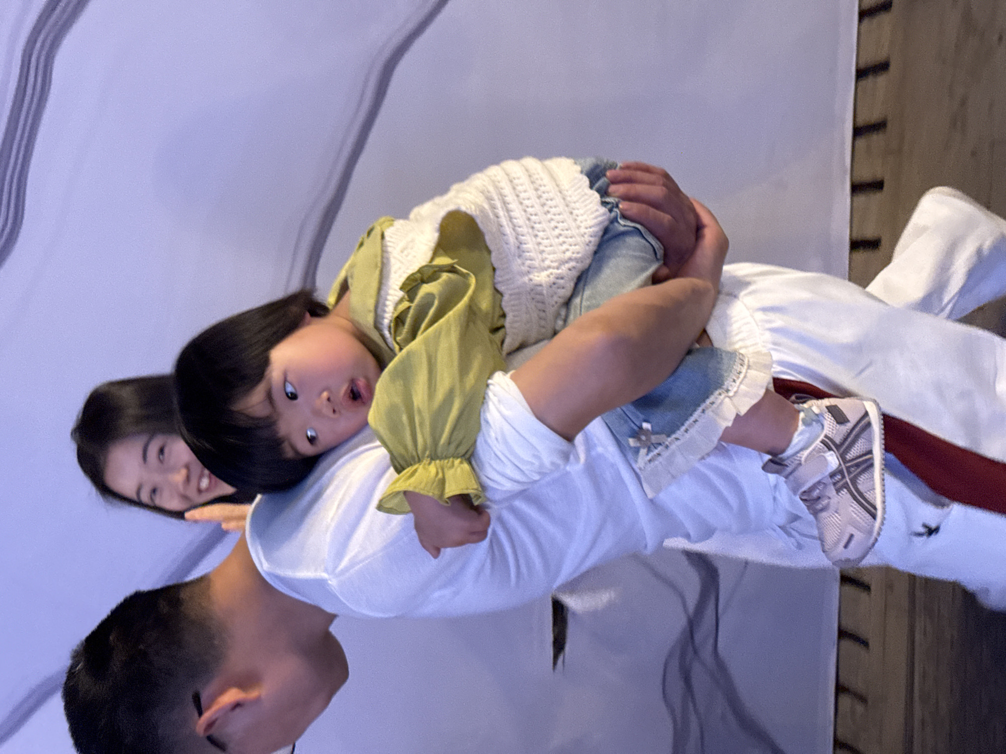 A family photograph depicts a man, a woman, and a child, possibly taken in a residential setting within the Yuzhong District of China, based on the observable elements. The man carries the child on his shoulders, while the woman stands nearby. The child wears a green top, a white knitted vest, and jeans. The man wears a white t-shirt. The woman's attire is not clearly visible. The background includes striped decor and wooden accents. The child is looking forward with an excited expression.