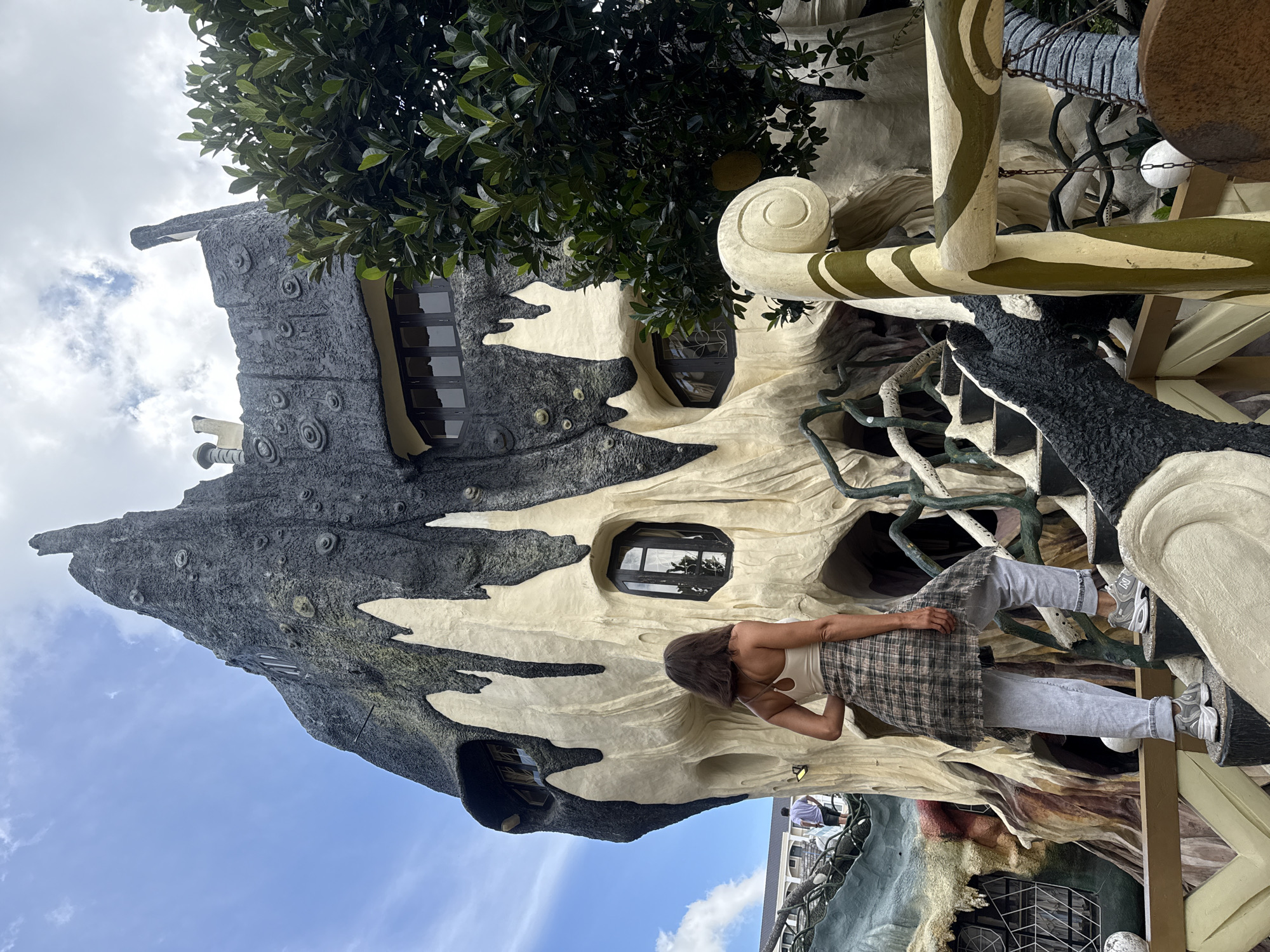 The image shows a woman standing on a staircase in front of a unique, organic-shaped building. The building appears to be constructed of concrete and is textured to resemble a tree trunk, with dark gray, rough surfaces and lighter, smoother sections that look like dripping wax or sap. There are several irregularly shaped windows that resemble eyes, and the overall structure has an almost creature-like appearance. The woman is wearing a light-colored tank top, a plaid shirt tied around her waist, light-colored jeans, and athletic shoes. She is facing away from the camera, looking up at the building. The staircase she is on is also irregularly shaped and blends with the architectural style of the building. In the background, there are trees and a cloudy sky. This building is known as the Hang Nga Guesthouse, also commonly referred to as the "Crazy House," located in the city of Da Lat, Vietnam.