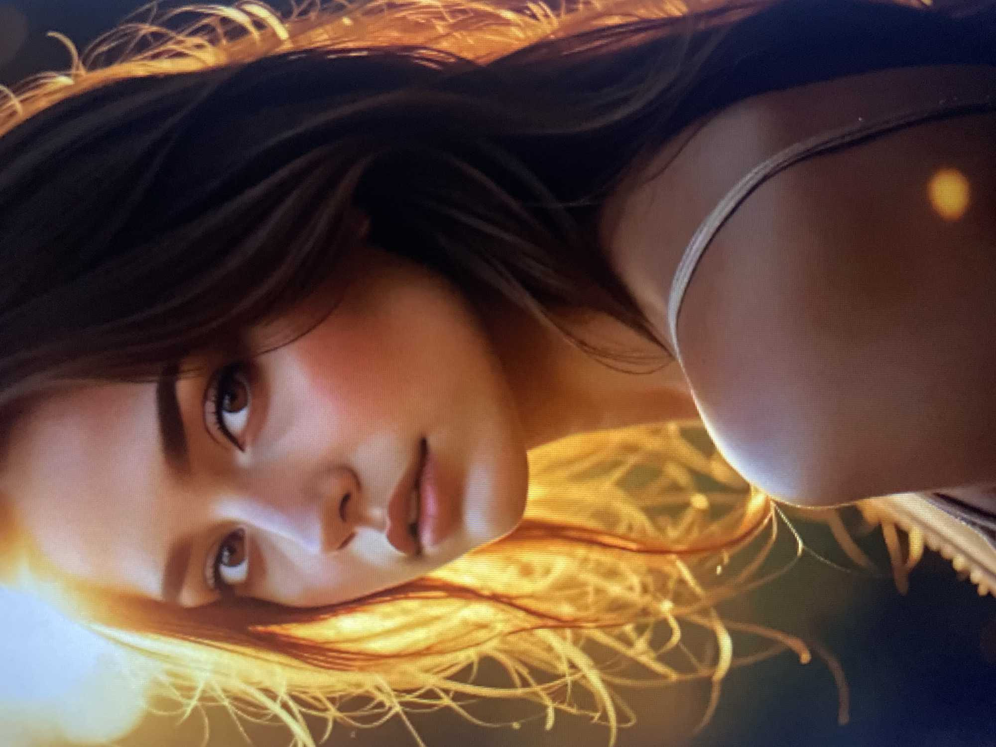 The image shows a close-up portrait of a young woman with long, dark brown hair, illuminated by warm, golden light that appears to be sunlight. She is looking towards the viewer with a soft expression. Her skin is smooth and her makeup is subtle, with a touch of blush on her cheeks. She is wearing a thin strap, suggesting a tank top or dress. The background is blurred, with bokeh effects from the light source, creating a dreamy and atmospheric feel. The scene is not set in any specific city or location; it is a generic portrait.