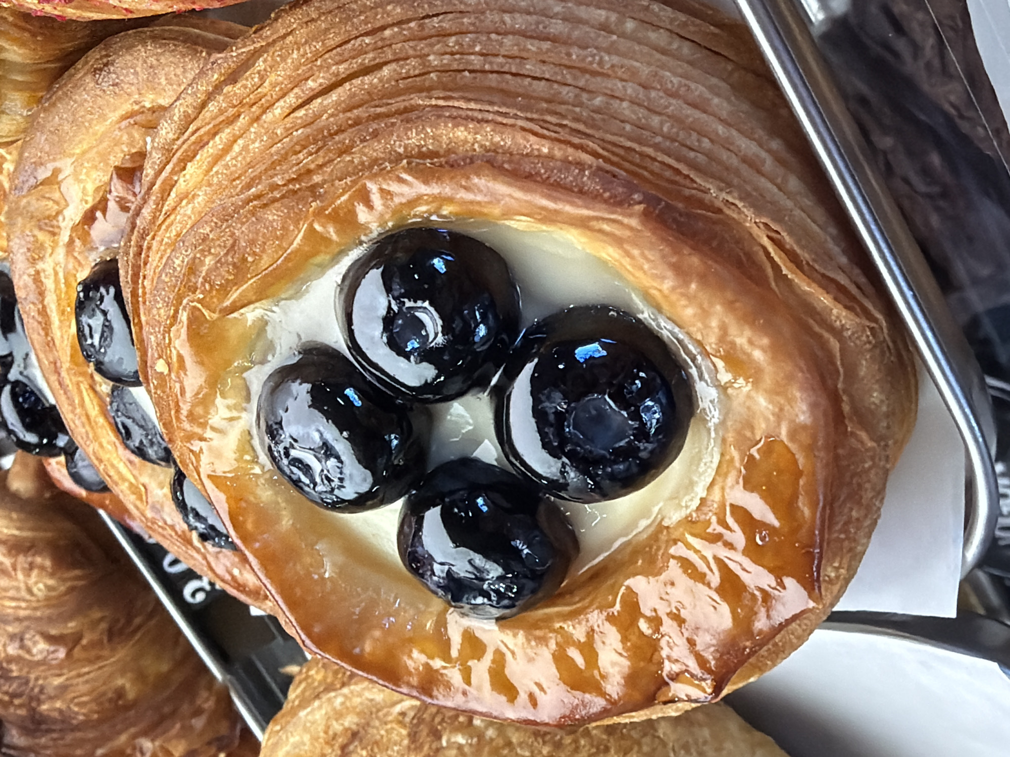 The image shows a close-up of a glazed pastry with a filling of cream and dark, shiny berries (likely cherries). The pastry is golden brown and appears flaky. It is sitting on a tray or display, possibly in a bakery or café. There is no information related to the location of the city of Izhevsk, Russia, in the image. There are no people visible.