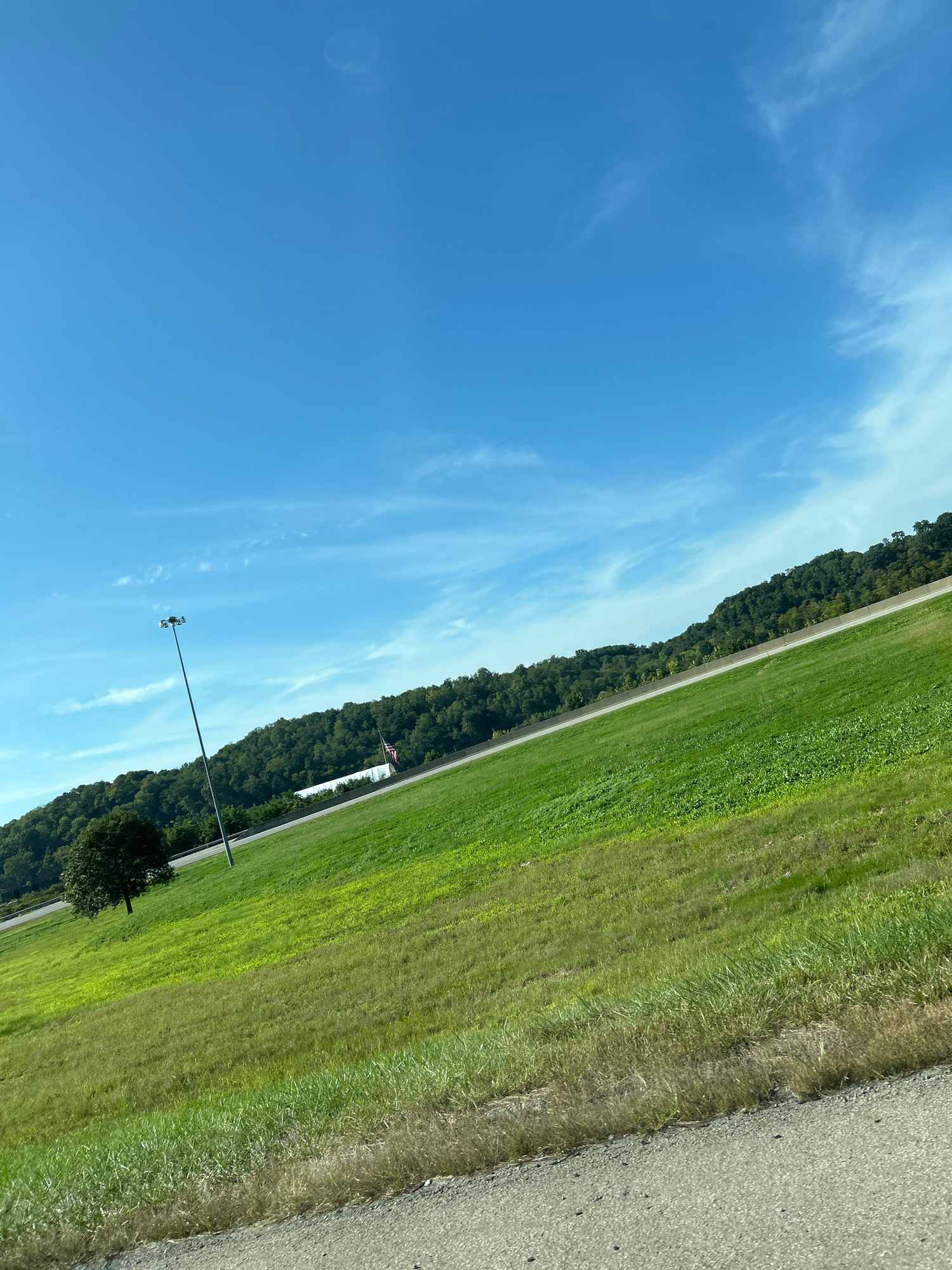 The image depicts a roadside scene on a clear, sunny day in Collier Township, Pennsylvania, United States of America. A bright blue sky with sparse, wispy clouds dominates the upper portion of the frame. Below the sky, a gently sloping, vibrant green grassy hill stretches across the image. A tall, thin lamppost stands on the left side of the hill, its light fixture clearly visible. In the middle ground, a paved road runs parallel to the base of a densely wooded hillside. An American flag is visible on a flagpole near the road, alongside a white building. The foreground shows a portion of a textured gray asphalt surface, likely a road or shoulder, with some dry grass along its edge. The overall impression is one of a rural or suburban landscape under a bright sky.
