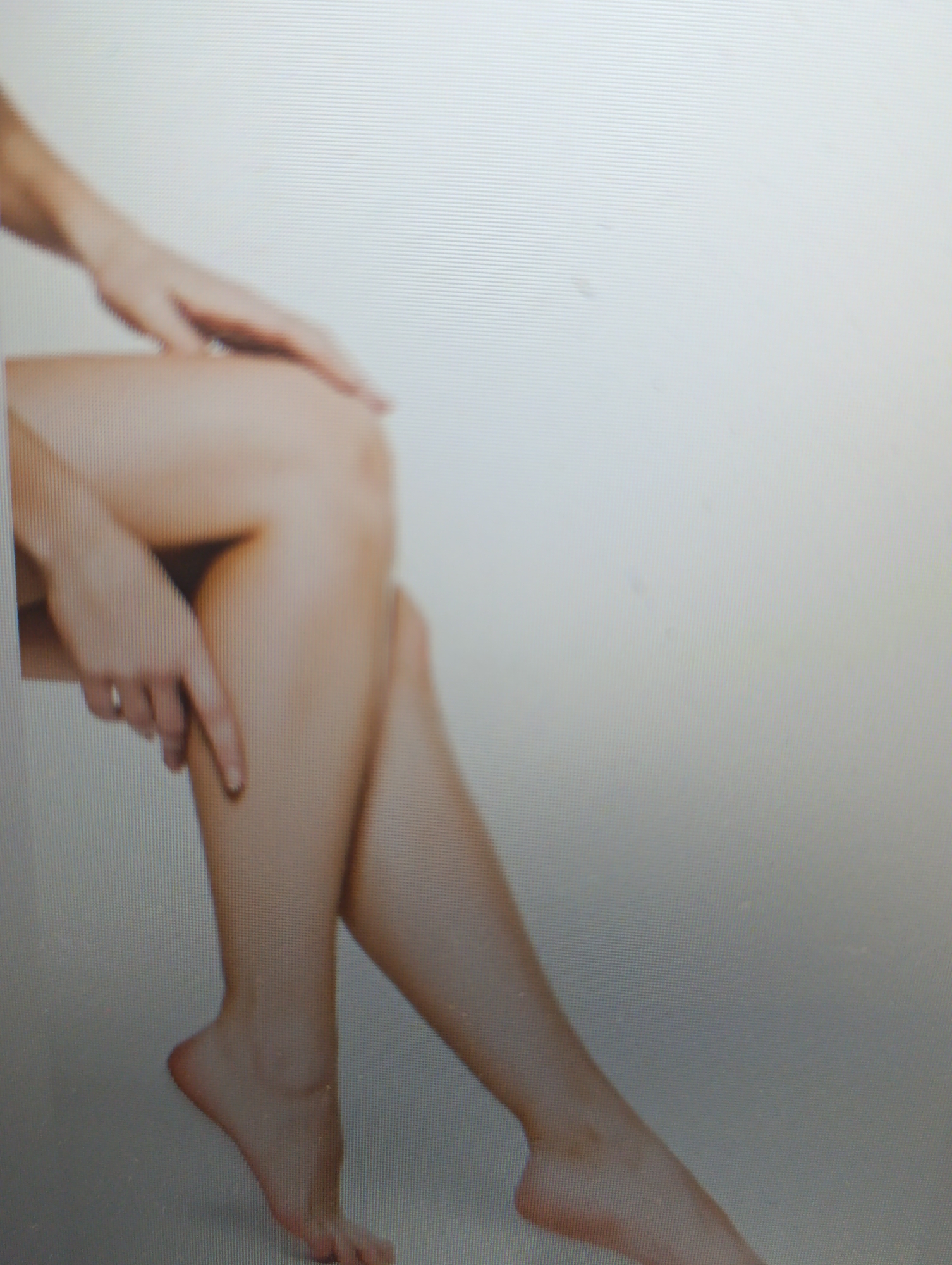 A full body shot of a person's legs is shown, taken from a low angle. The person's skin is fair and smooth. Their left hand is resting on their right thigh. The person is seated with their legs crossed. Their feet are bare and visible. The background is a light-colored wall with horizontal lines. The image appears to be taken indoors, with soft lighting.