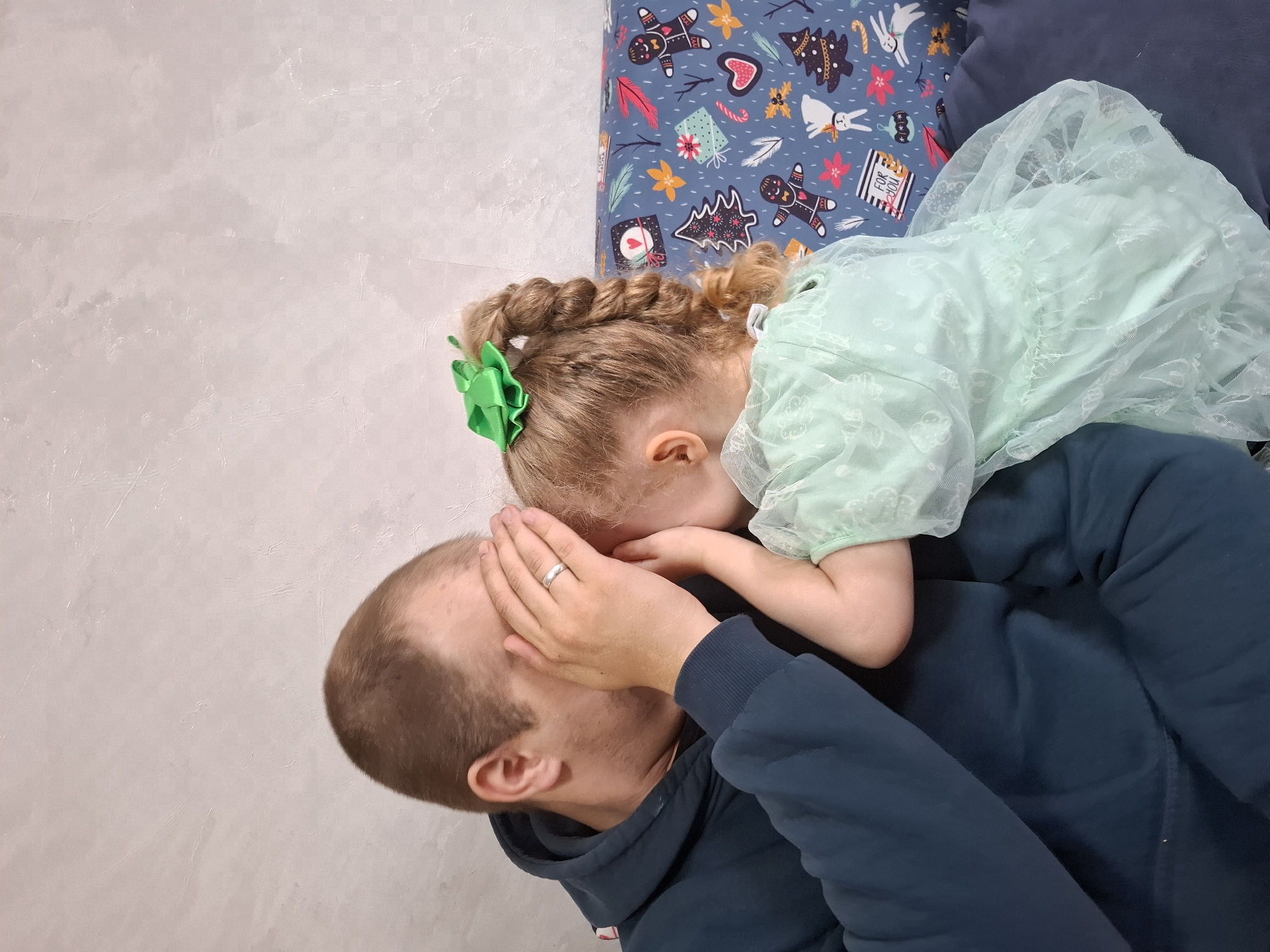 A man and a young girl are lying on what appears to be a sofa or bed. The man, who has a shaved head, is wearing a dark blue hooded sweatshirt and has his hand covering his face. A wedding band is visible on his ring finger. The young girl, wearing a light green dress and a green bow in her braided hair, is leaning in towards the man. The bedding beneath them is patterned with Christmas-themed designs, including gingerbread men, Christmas trees, and other holiday motifs. The background includes a plain wall with a subtle texture. The image was taken in the city of Chaikovsky, Russia.