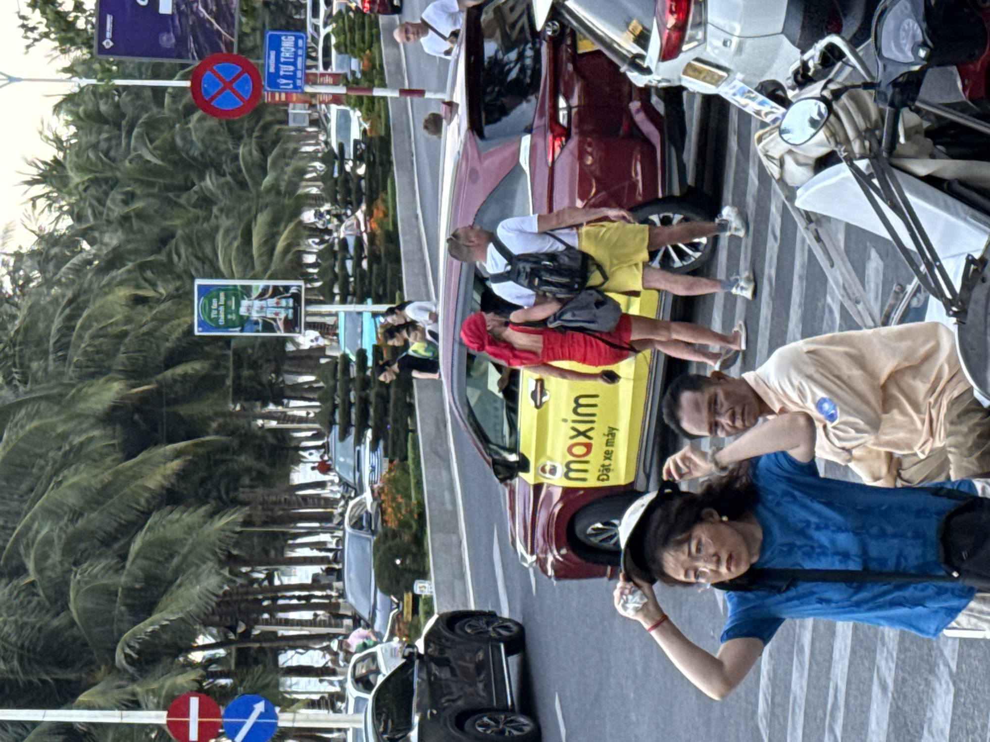 The image captures a street scene in Nha Trang Ward, Vietnam, focusing on a crosswalk. In the foreground, a woman in a blue t-shirt and glasses is adjusting a helmet on her head. Beside her, a man in a light tan shirt sits. Behind them, a yellow and burgundy Maxim car, a ride-hailing service, is stopped. A woman in a red dress and a man in yellow shorts are walking away from the car. In the background, palm trees line the street, and several cars are visible, suggesting a busy urban environment. Traffic signs are present, including a "No Parking" sign. The lighting suggests it is daytime.