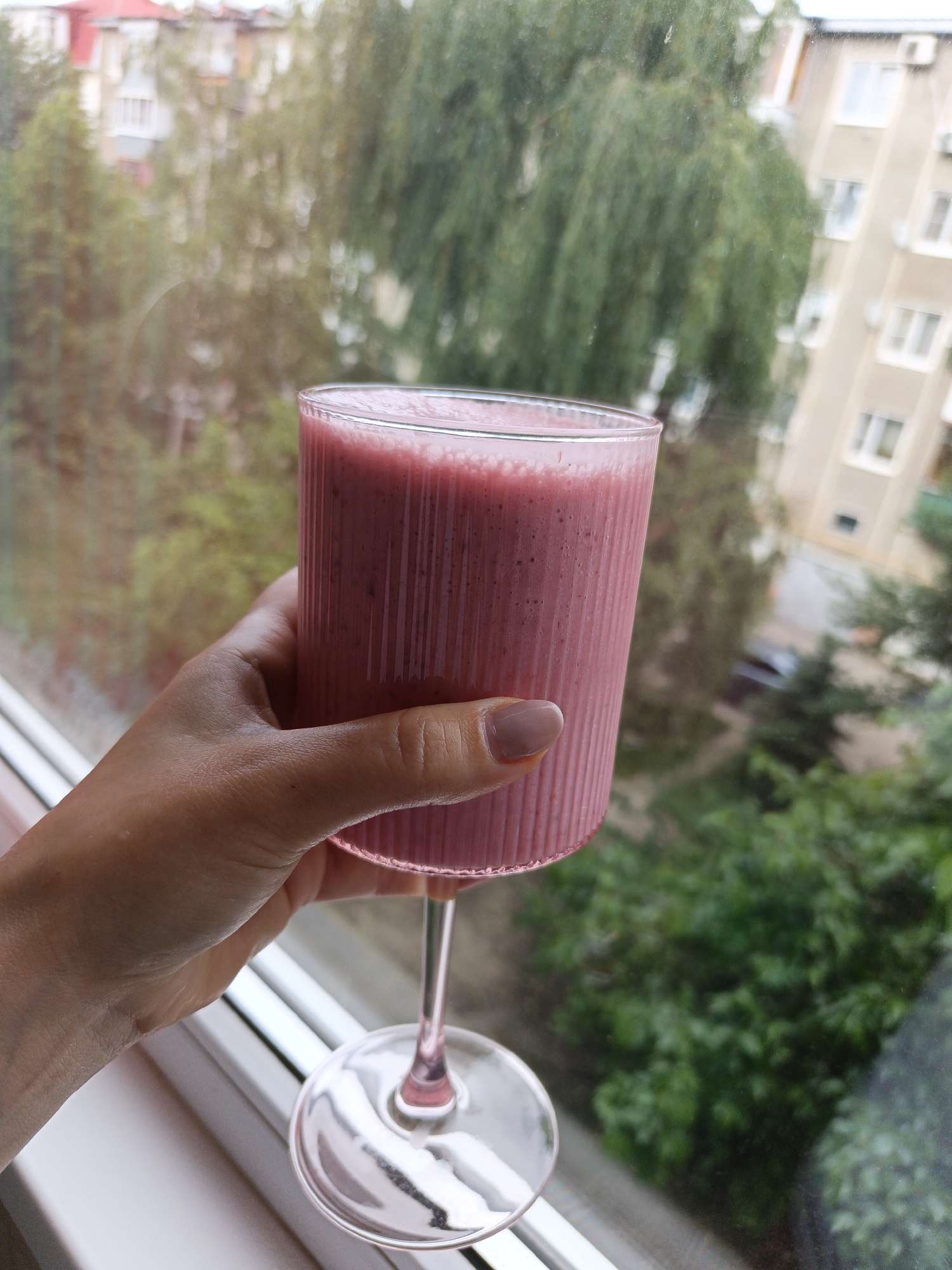 A woman's hand is holding a glass of pink smoothie in front of a window. The window looks out onto a scene of trees, buildings, and the city. The glass is ribbed and on a stemmed base. The person has light skin tone with neutral color nail polish. The background appears to be in Goryachiy Klyuch, Russia.