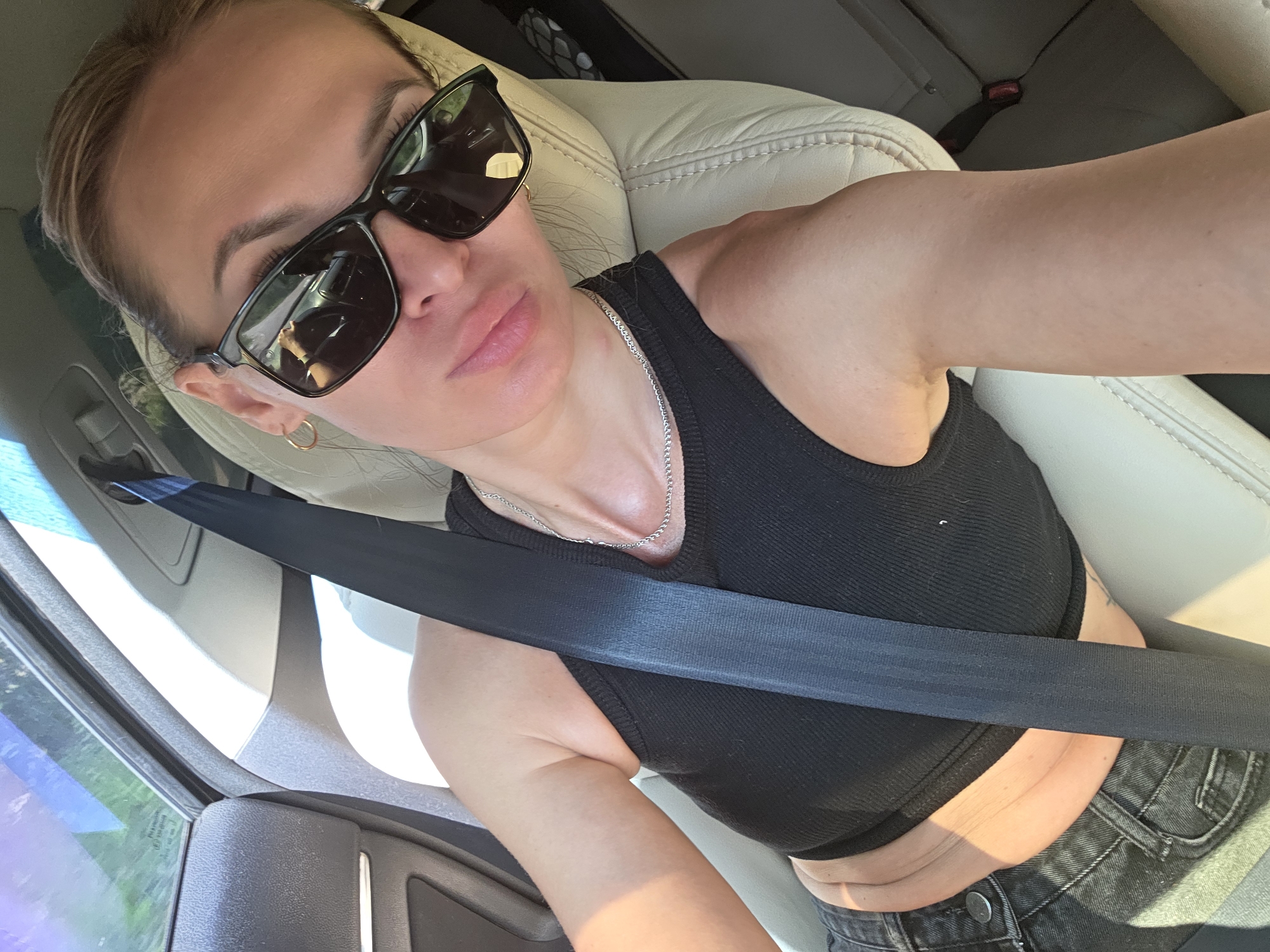 A woman with blonde hair pulled back is sitting in the passenger seat of a car, wearing sunglasses, a black tank top, and black jeans. A black seatbelt is fastened across her chest. She is taking a selfie with her phone. The car's interior, including a beige leather seat and parts of the dashboard and door panel, is visible. The scene appears to be outdoors, with sunlight filtering through the car windows, suggesting a daytime setting. There is no discernible urban or natural landmark to indicate a specific location in Noviy, Russia.