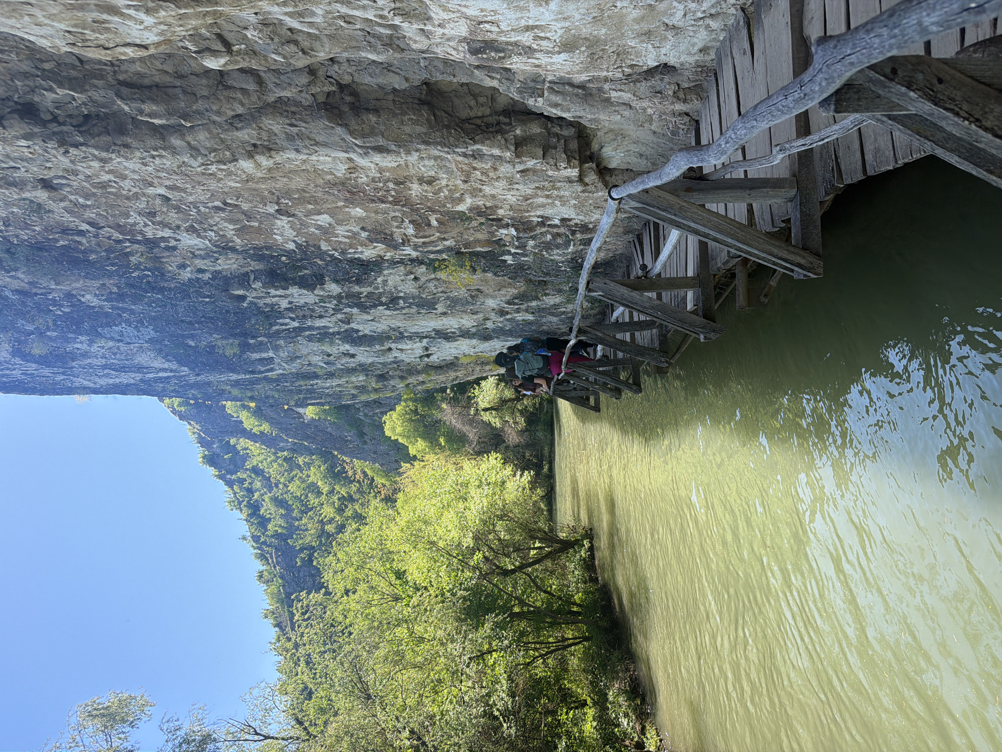 A low-angle, slightly tilted landscape photograph depicts a wooden bridge clinging to a rock face over a river. The bridge, made of weathered wood, has an irregular construction with support beams extending into the murky water. Two figures are on the bridge, one wearing a red shirt and the other in green. Their activities are not discernible. Above the bridge, a rock face reveals uneven terrain and slight vegetation growth. Lush green trees line the lower left of the frame. The sky appears clear, suggesting daylight. This scene is located at Petrevene, Bulgaria.