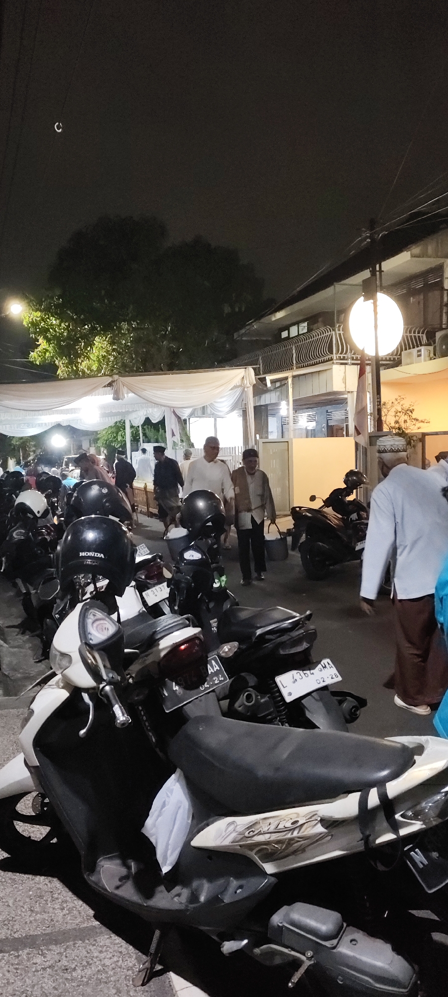 The image captures a nighttime scene in a city, likely in Malang, Indonesia, as indicated by the Indonesian license plates visible on the motorcycles. In the foreground, several parked motorcycles are prominent, some with helmets placed on them. The lighting suggests an urban setting with streetlights illuminating the area.
In the background, a group of people are walking, some of whom appear to be wearing traditional religious attire, indicating a possible religious gathering or event. A white tent or canopy is set up, suggesting an outdoor event. A building with a balcony is visible to the right, with architectural details common in Indonesian cities. The presence of a street lamp and overhead wires further confirms an urban environment. The overall atmosphere is one of a community event taking place after dark.