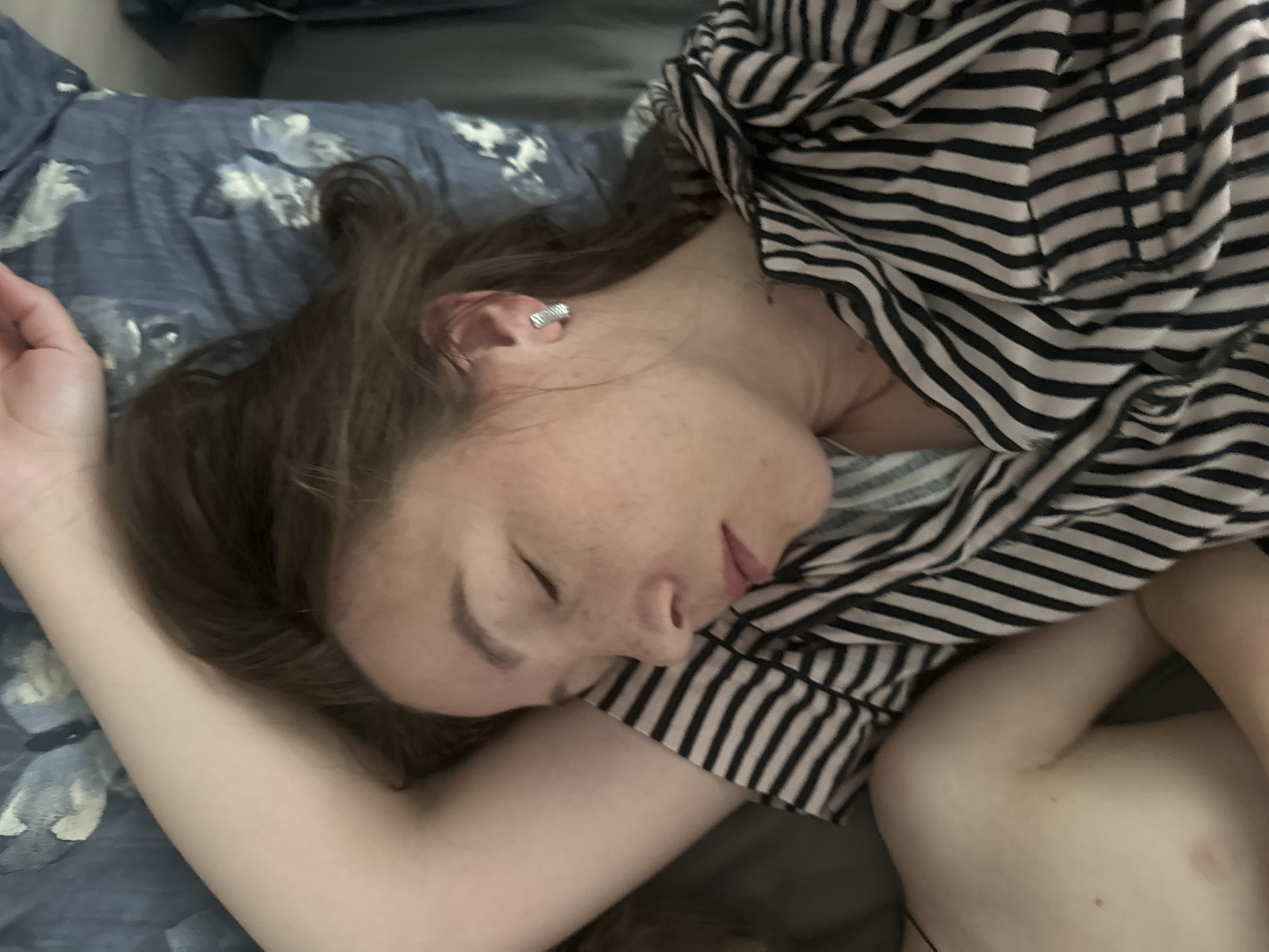 A woman is sleeping on a bed. She has brown hair, fair skin, and is wearing a striped top. The bed has a floral patterned sheet and a grey pillow. The location is Chaikovsky, Russia.