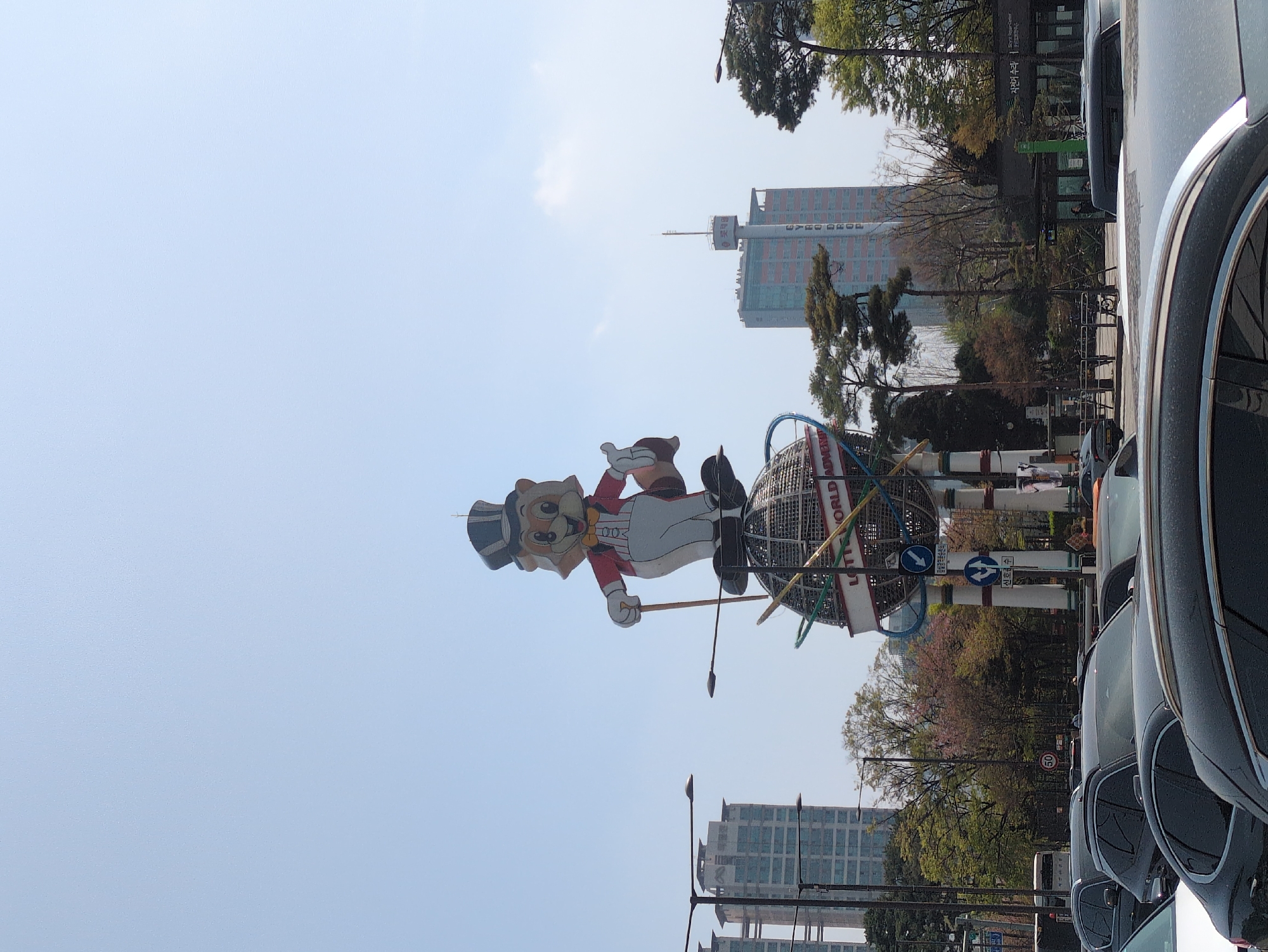 The image shows a large, colorful cartoon character display for Lotte World Adventure amusement park in Seoul, South Korea. The character, a bear in a top hat and red coat, stands atop a globe-like structure. Buildings are visible in the background. Cars line the street in the foreground. Signage in Korean script is also visible. The sky is light blue and appears clear.
