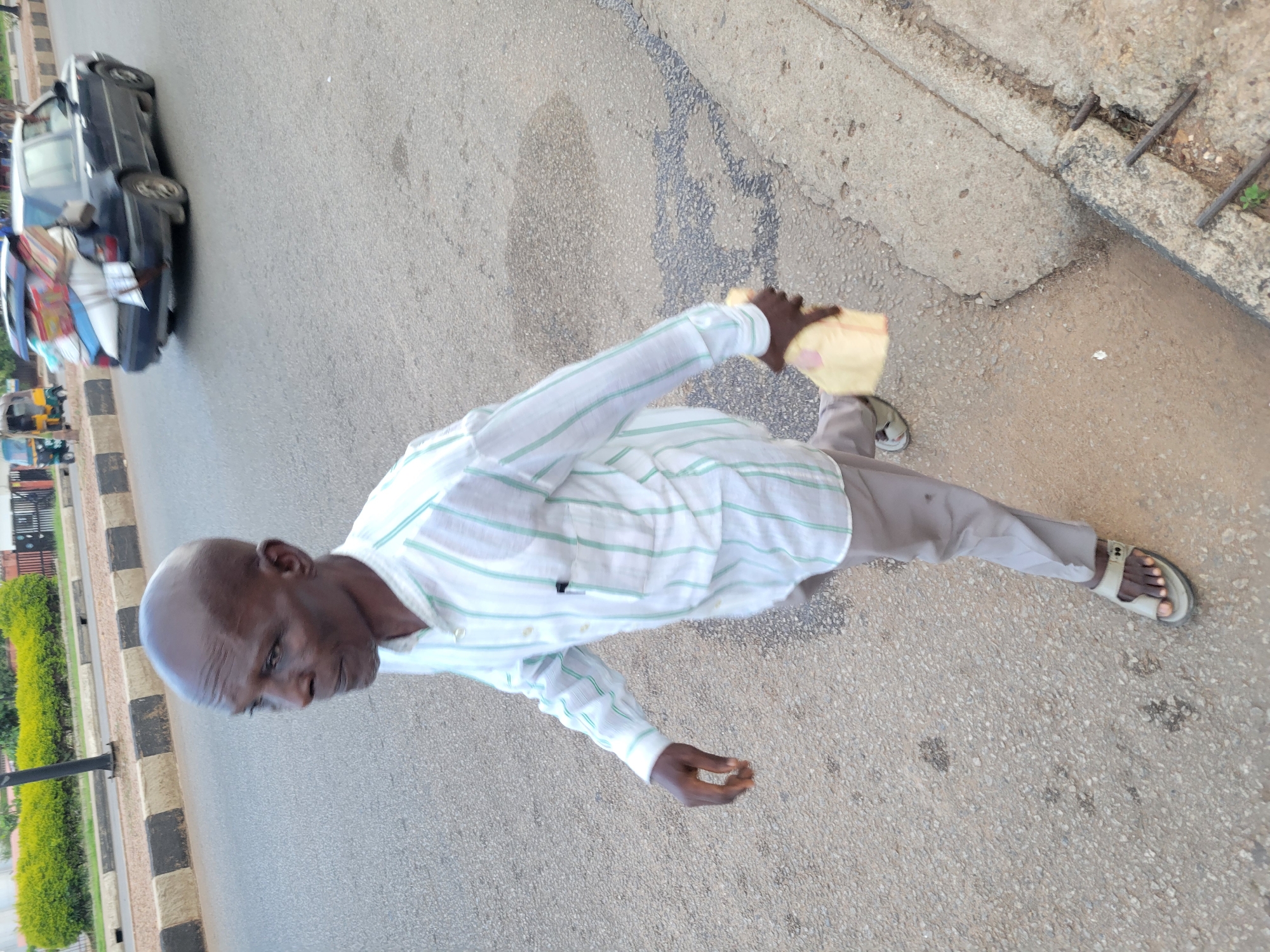 An elderly bald Black man wearing a white and green striped long-sleeved shirt and light gray pants walks on a paved road in Jos, Nigeria. He is carrying a light yellow package in his right hand. A dark blue sedan with its trunk open and filled with luggage is driving in the background. There are also yellow and blue tricycles on the road. The ground is asphalt and concrete, with some visible potholes and cracks. A concrete curb with exposed rebar is visible on the right side of the frame. Green foliage and a street lamp are visible in the background on the left.